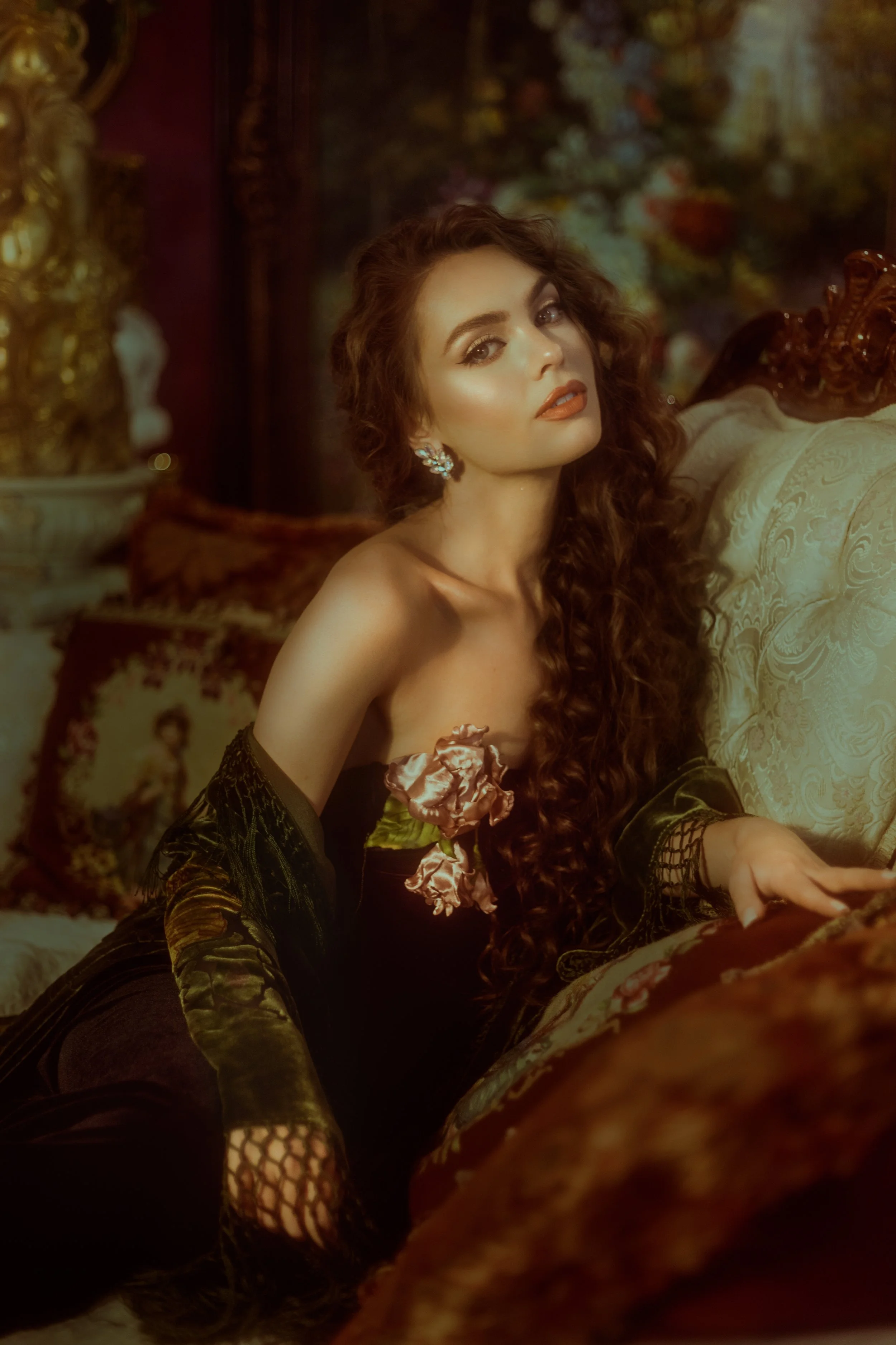 A woman with long, curly brown hair and wearing earrings, dressed in a black off-shoulder outfit with floral details, lying on a luxurious sofa in a vintage, ornate room with elaborate decor and artwork.