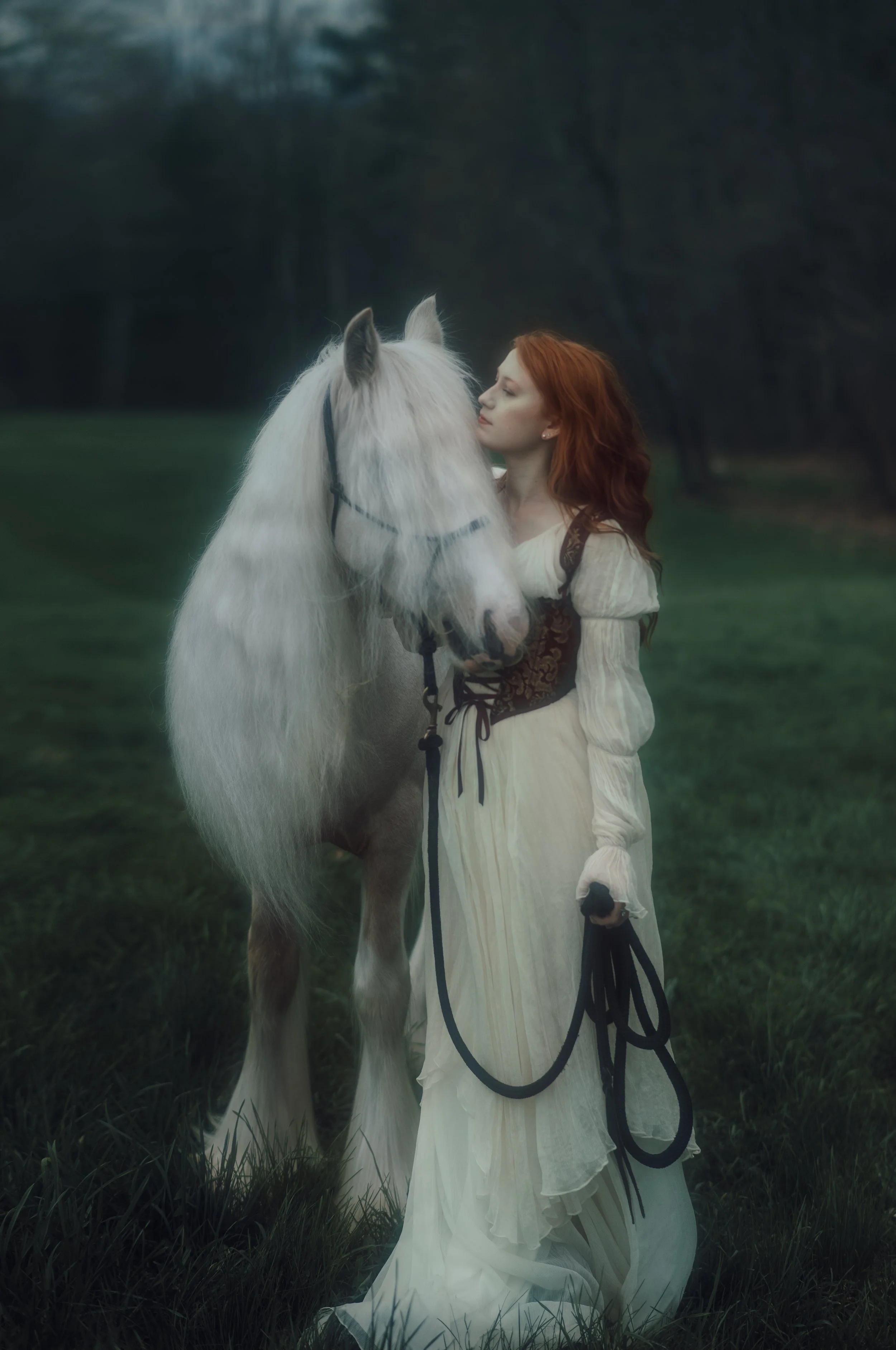 A woman with long red hair in a white vintage-style dress standing in a grassy field, gently kissing a white horse with a long, flowing mane.