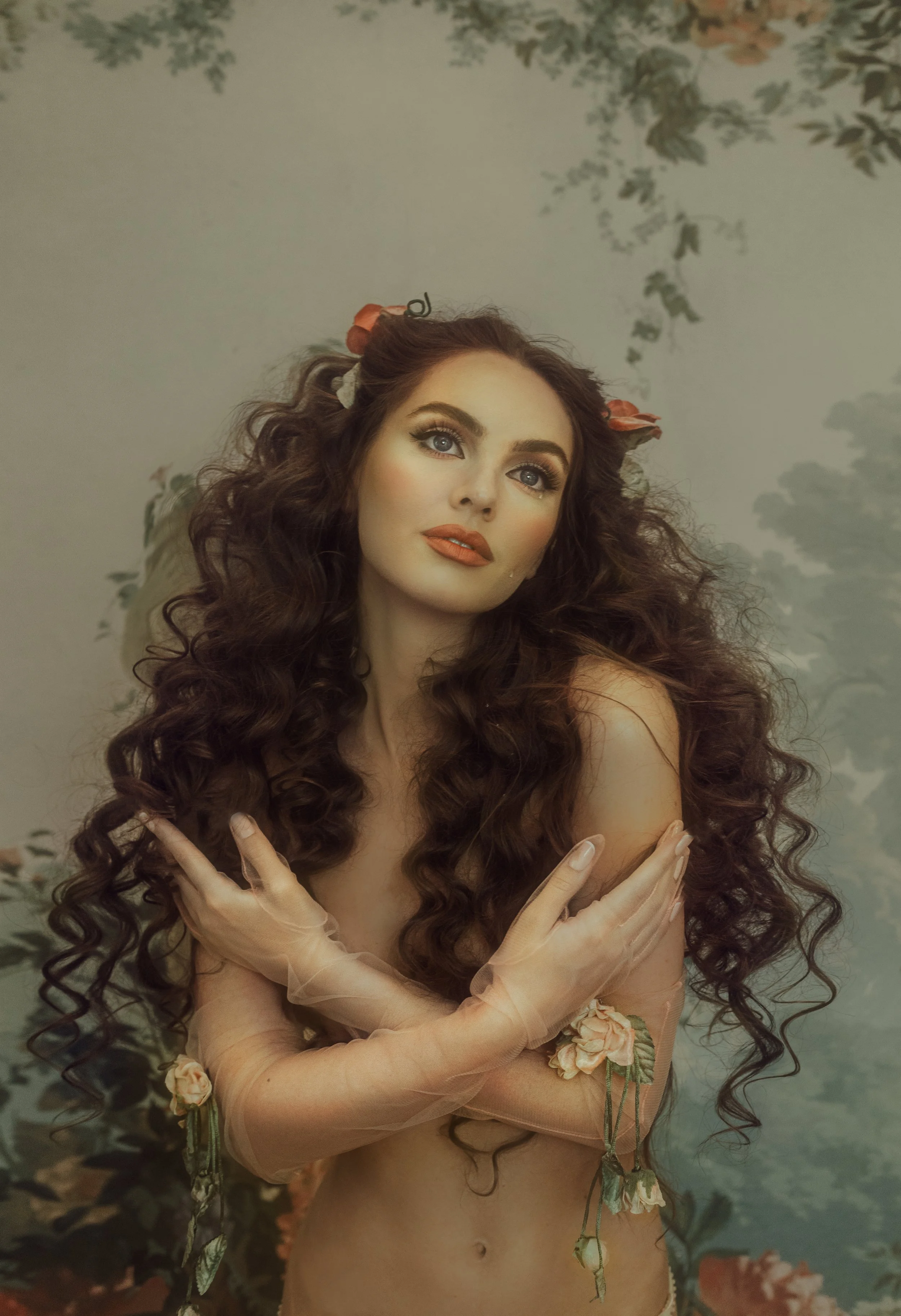 A woman with long, curly brown hair and floral hair accessories, wearing sheer peach-colored gloves with floral embellishments, posing with crossed arms in front of a painted nature background.