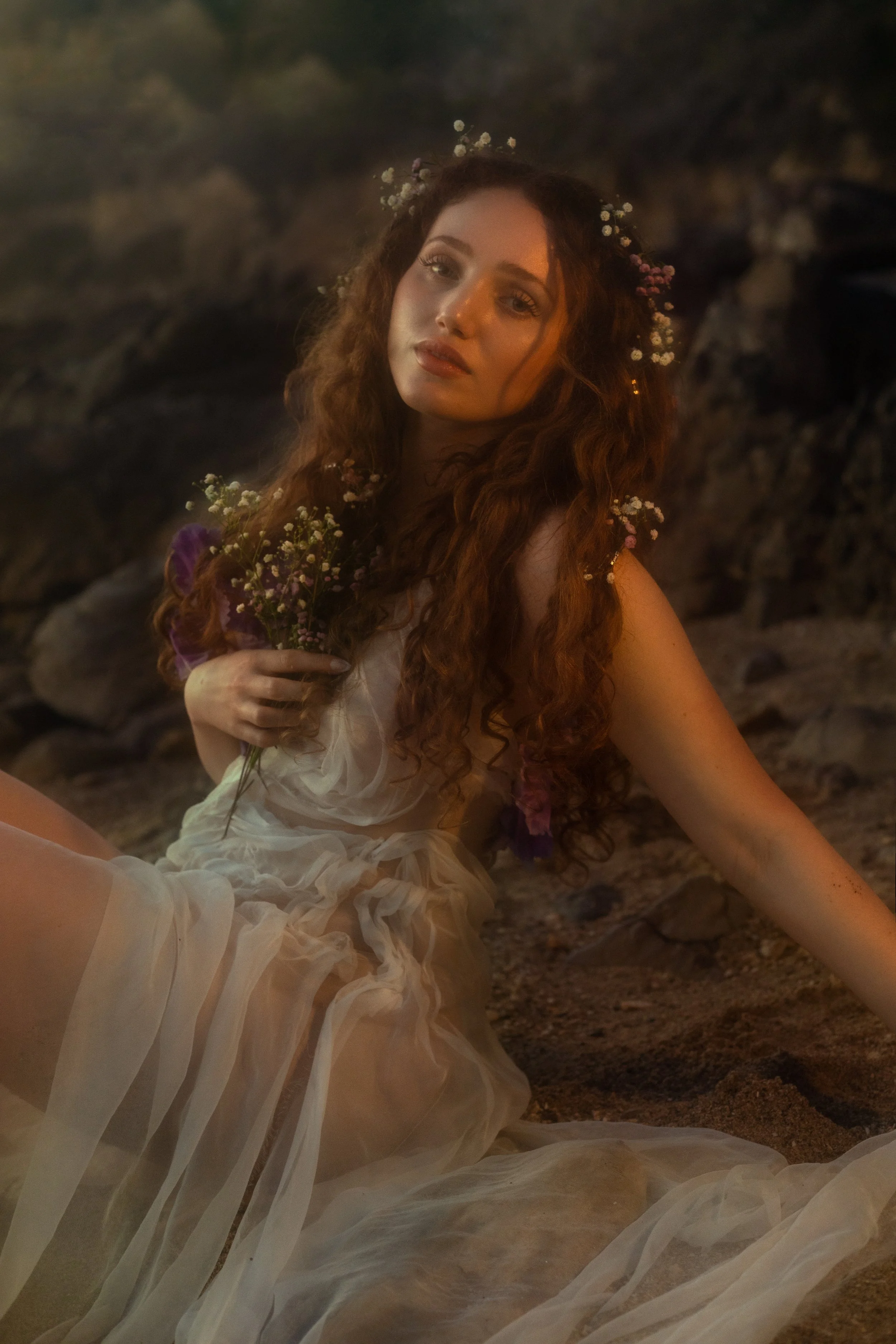 A woman with long, curly brown hair sits on a rocky ground in a dimly lit setting, wearing a flowing, sheer, off-white dress. She has flowers woven into her hair and holds a small bouquet of purple and white flowers. The background is dark and rocky.