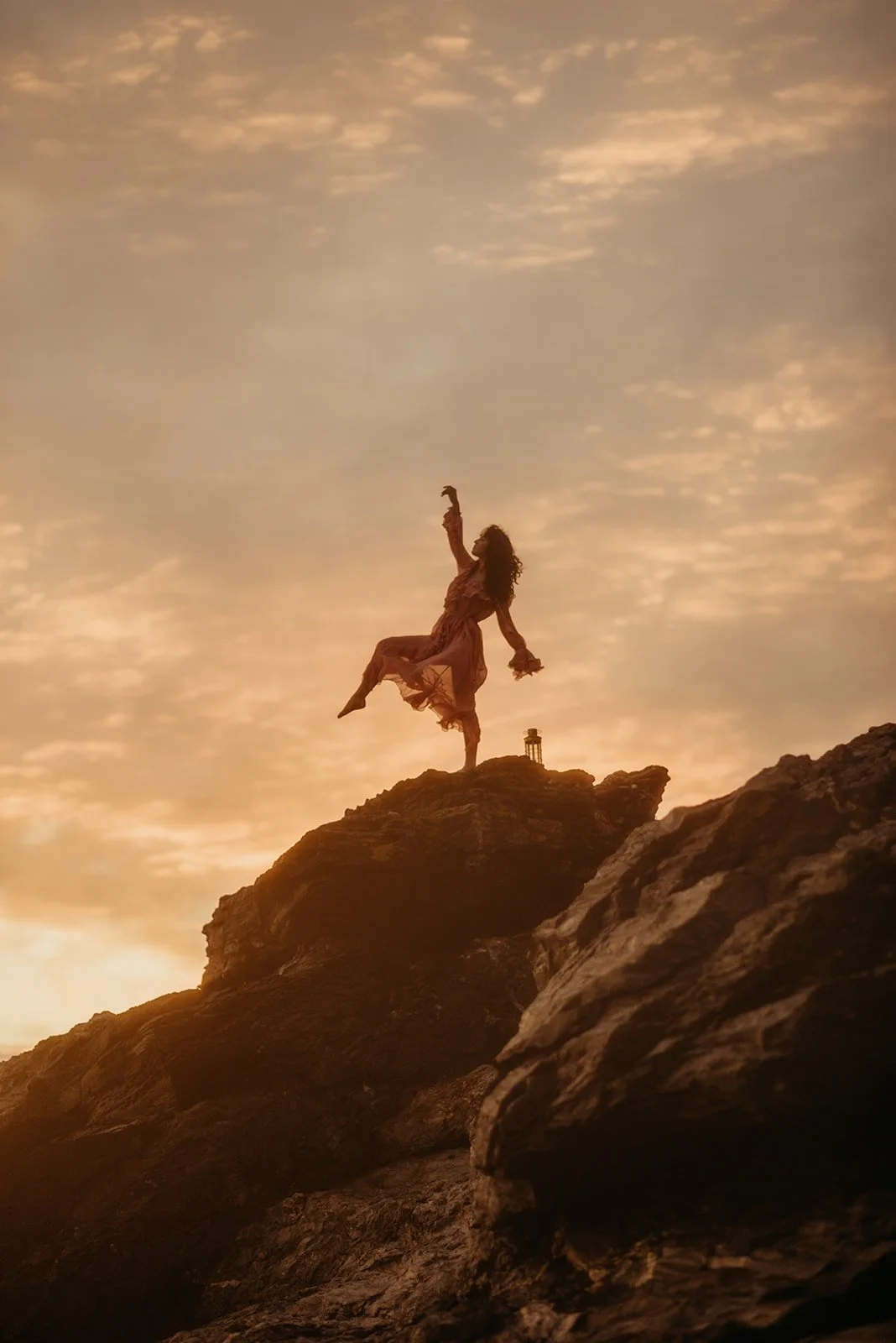 A woman in a flowing dress balances on one foot on a rocky hilltop during sunset, with her arms gracefully extended upward.