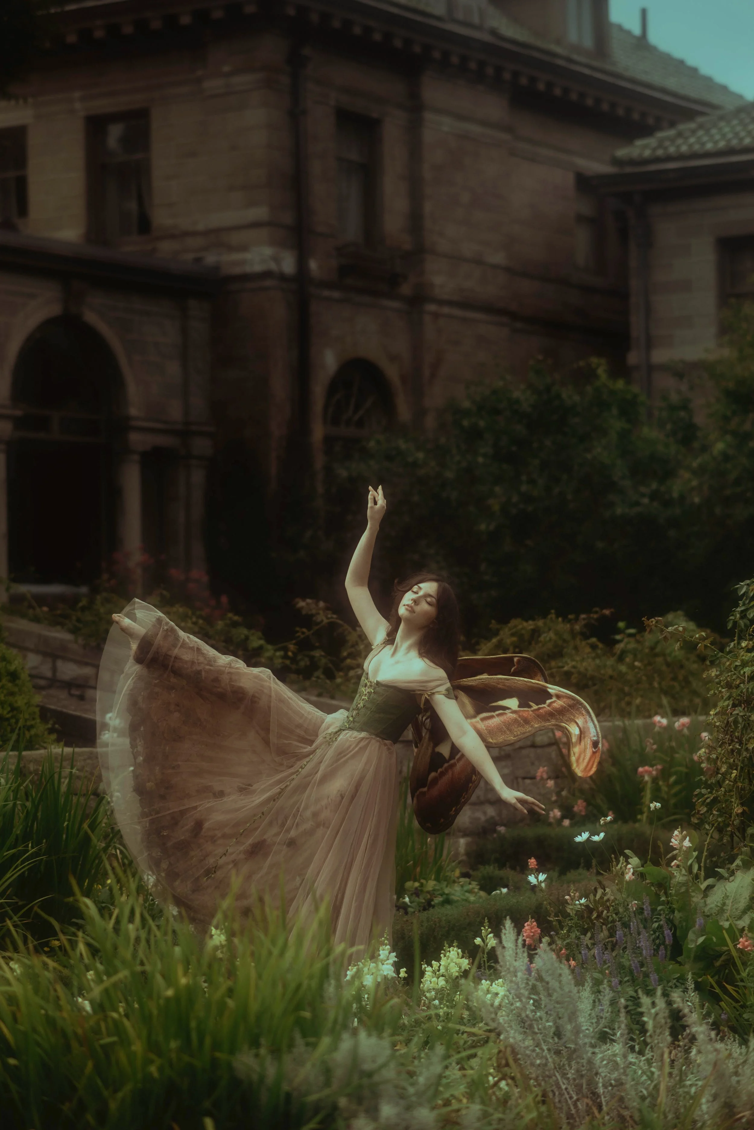 A woman in vintage dress poses in a garden with a butterfly on her arm, with an old stone building in the background.