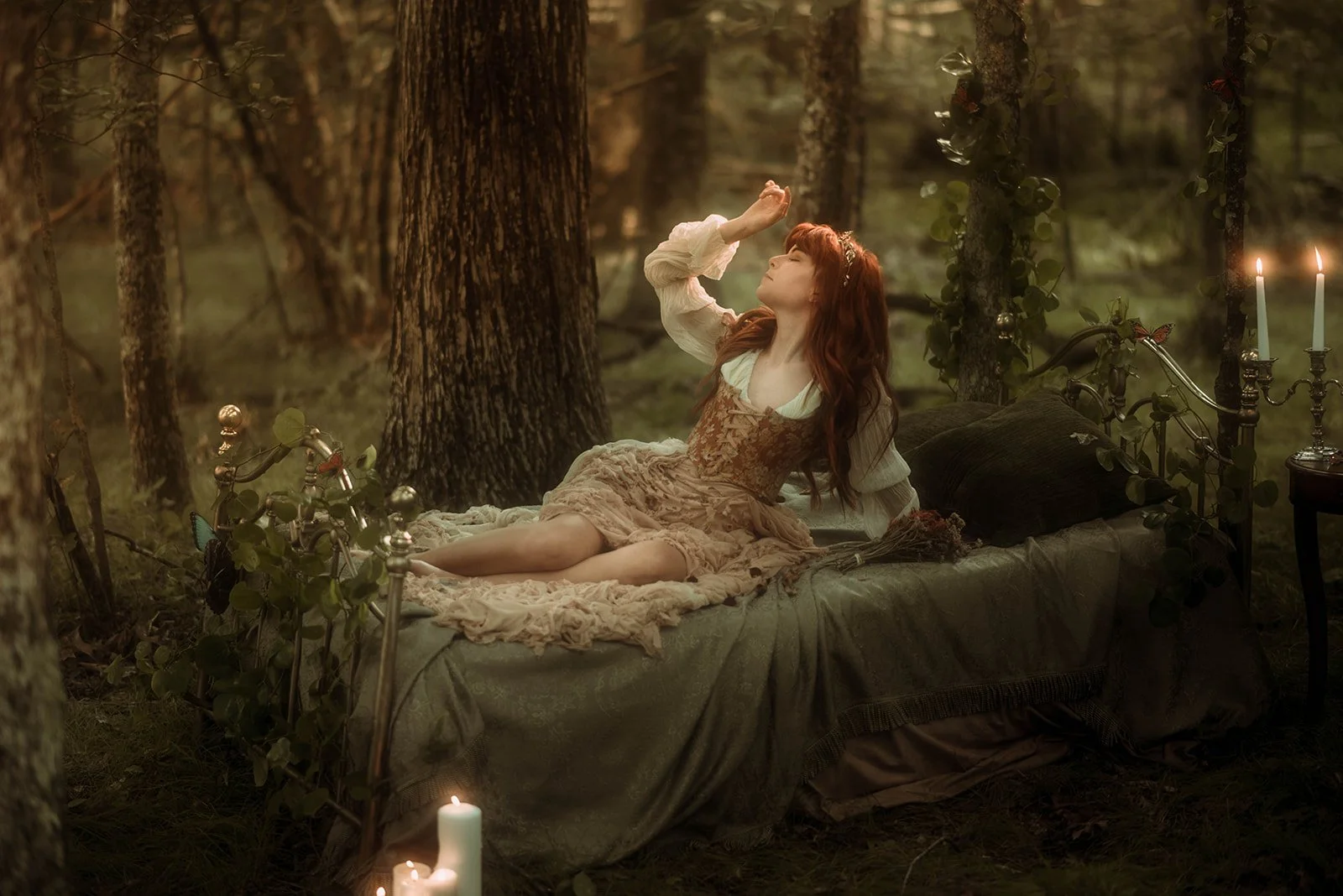 A woman with long red hair in a vintage dress lying on a decorated bed in a forest at sunset, surrounded by candles and butterflies.