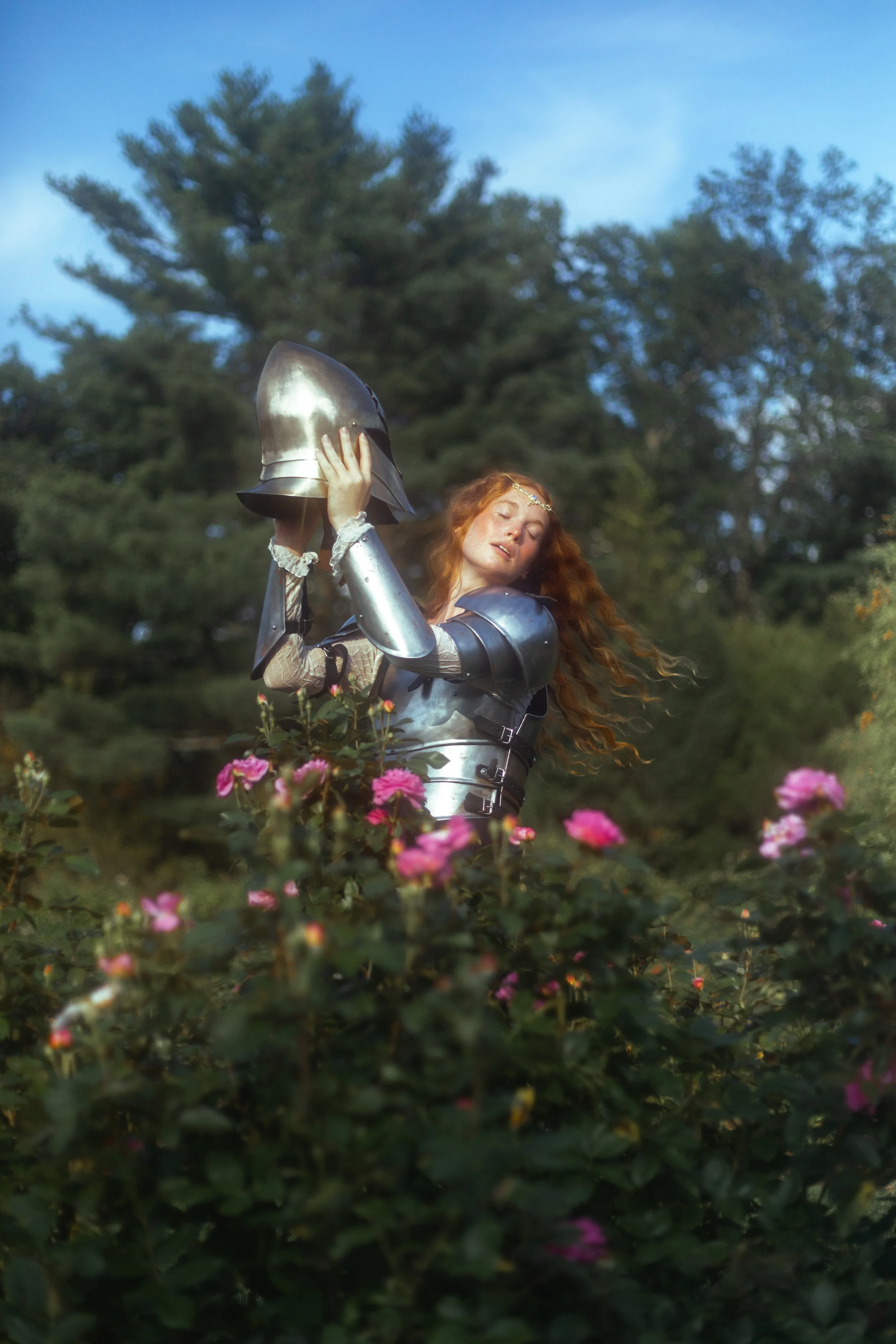 A woman dressed in medieval armor, holding a helmet above her head, standing among pink flowers with a backdrop of green trees and a blue sky.