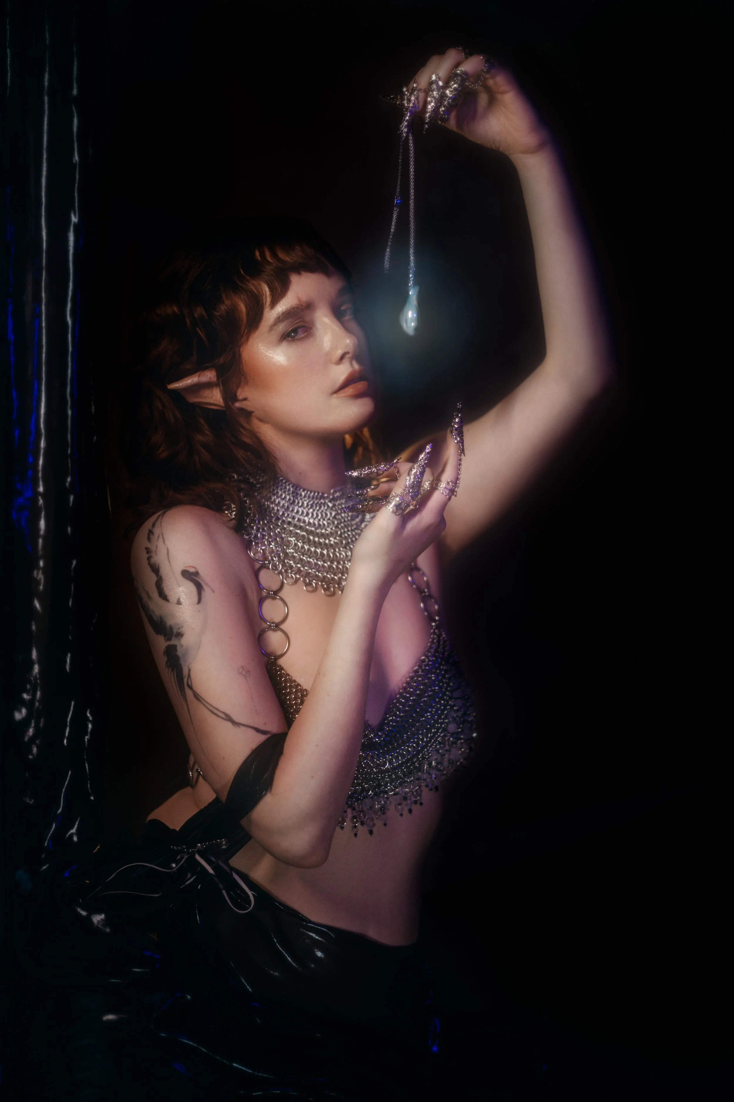 A woman with elf-like ears holding a necklace with a teardrop pendant in a dark setting, wearing metallic jewelry and a black, shiny outfit, with tattoos on her arm and shoulder.