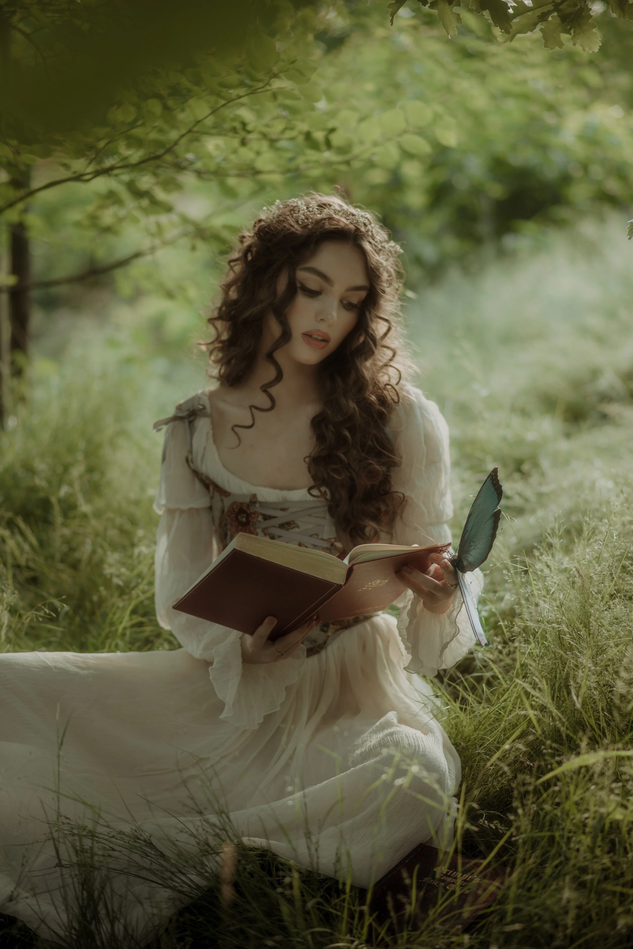 A woman with long, curly brown hair in a vintage white dress sitting in a lush, green outdoor setting, reading a book and having a butterfly land on her hand.