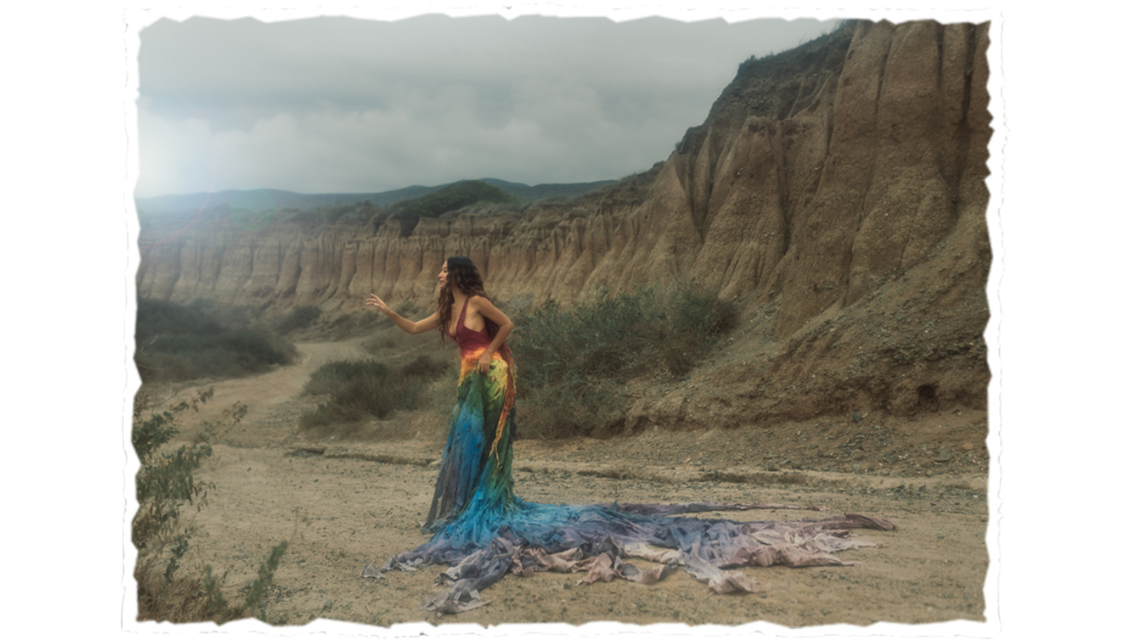 A woman with long curly hair wearing a rainbow-colored, long flowing dress standing in a desert with tall rocky cliffs in the background, reaching out with her right hand.