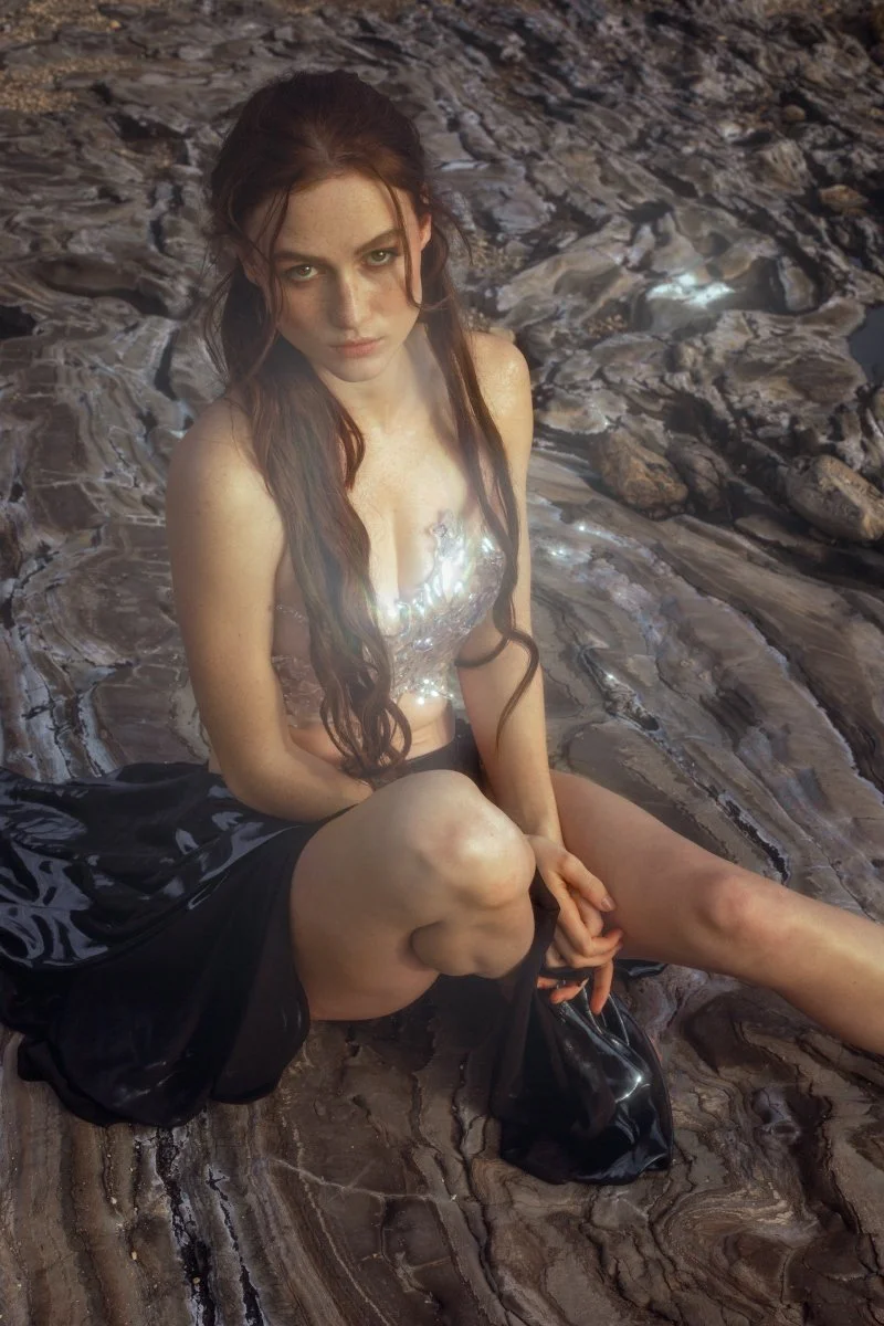 A woman with long dark hair and light skin sitting on a rocky surface, wearing a shiny, reflective top and black skirt.