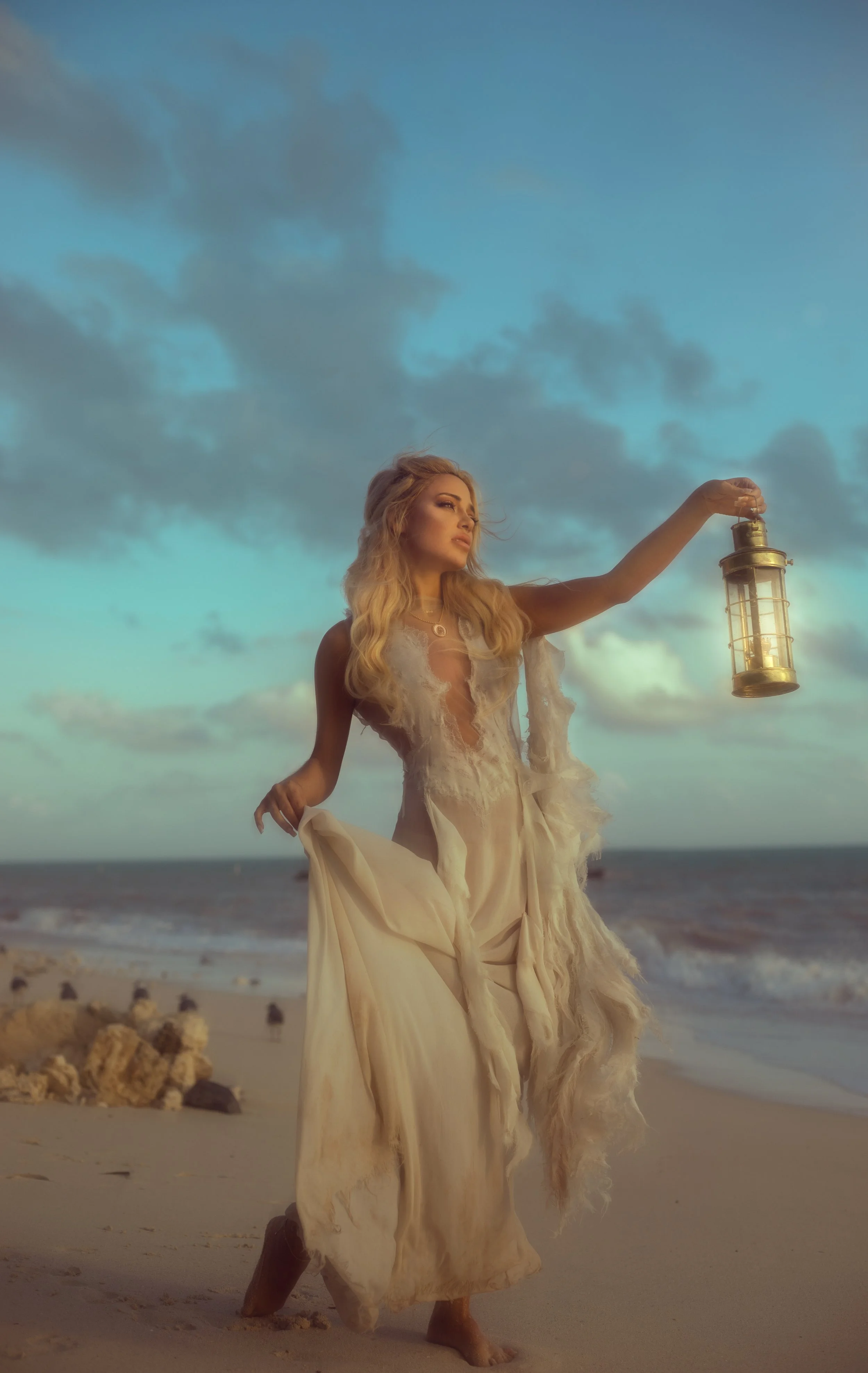 A woman in a flowing, distressed cream dress holds a lantern while standing barefoot on a beach with rocks, under a partly cloudy sky at sunset.