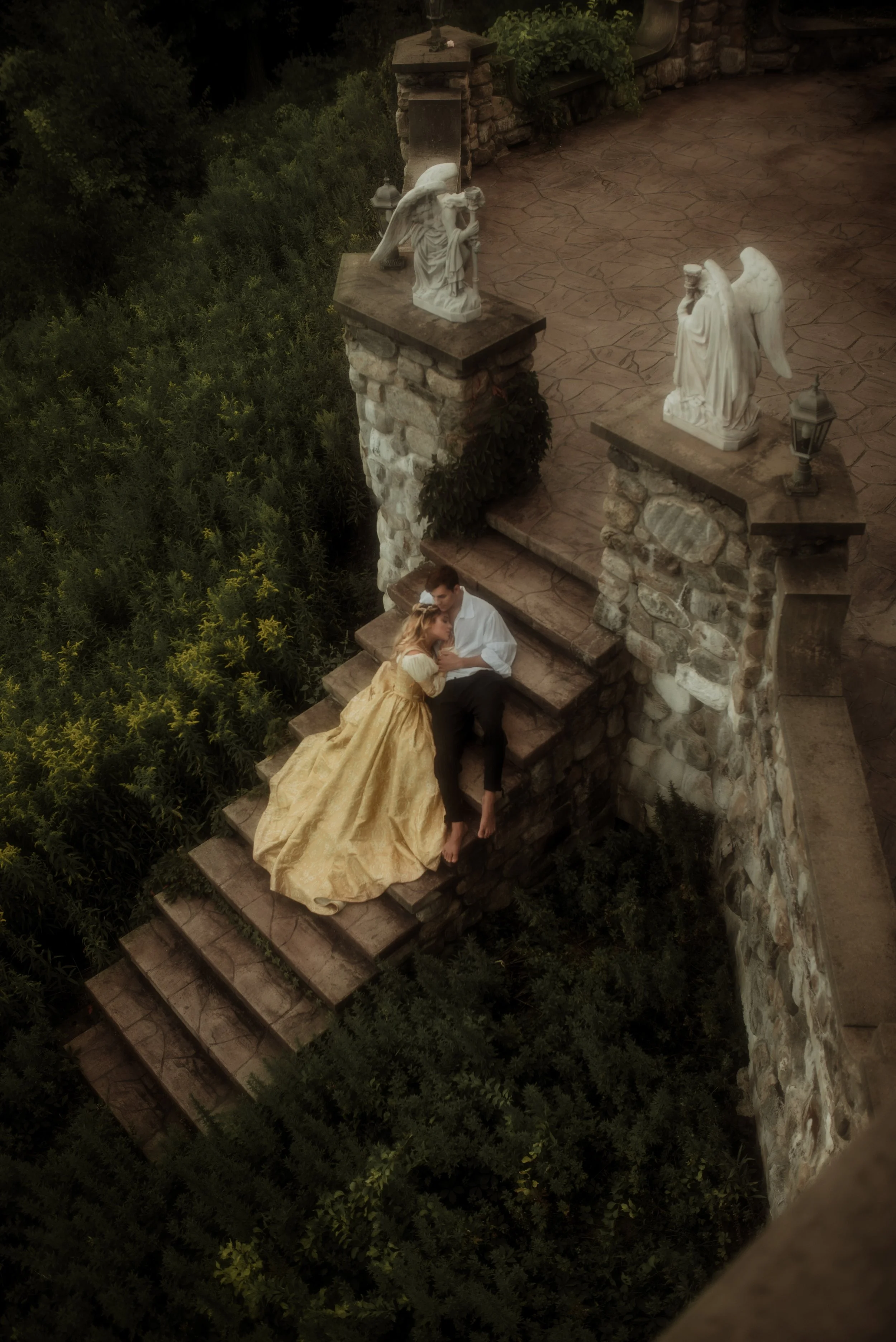 A romantic scene on stone steps features a woman in a long gold gown and a man in a white shirt and black pants sitting together. The woman is leaning on the man's shoulder, and they are holding hands. The background includes stone architecture, sculptures of angels, and lush greenery.
