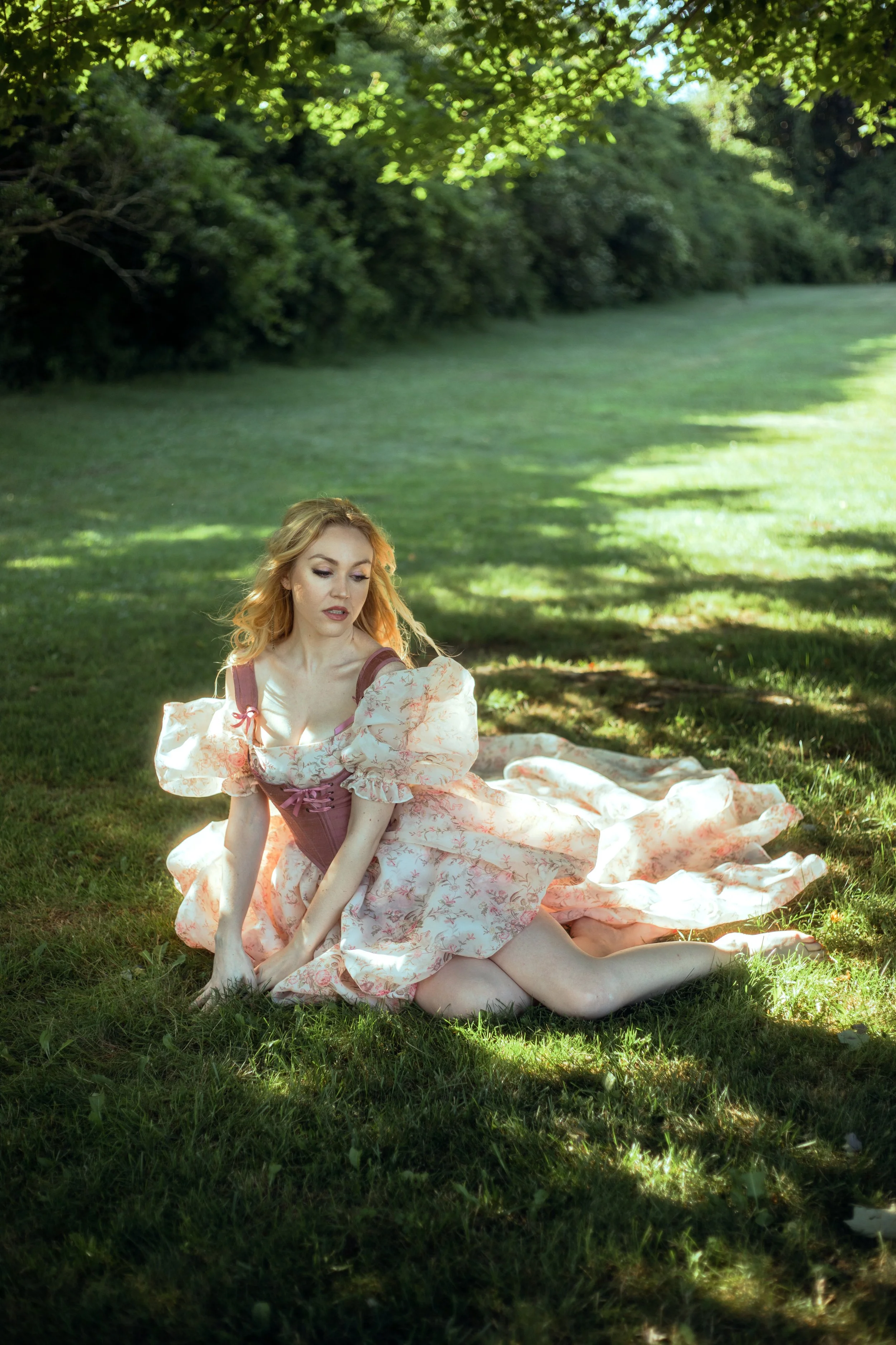 A woman in a vintage pink dress with puffed sleeves sitting on the grass in a park with lush green trees and sunlight filtering through.