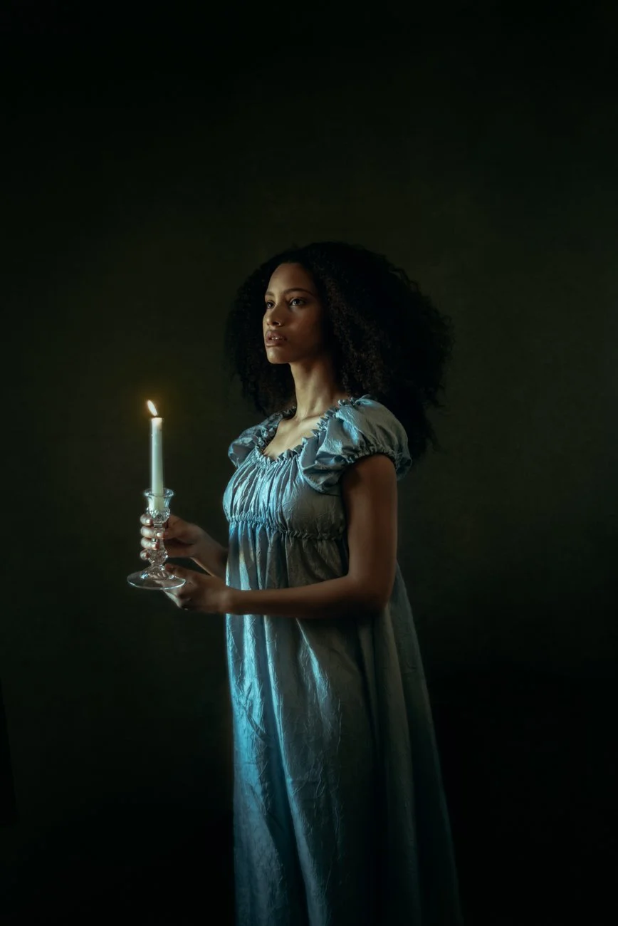 A woman with curly hair wearing a shiny, short-sleeved blue dress holding a lit candle in a candleholder against a dark background.