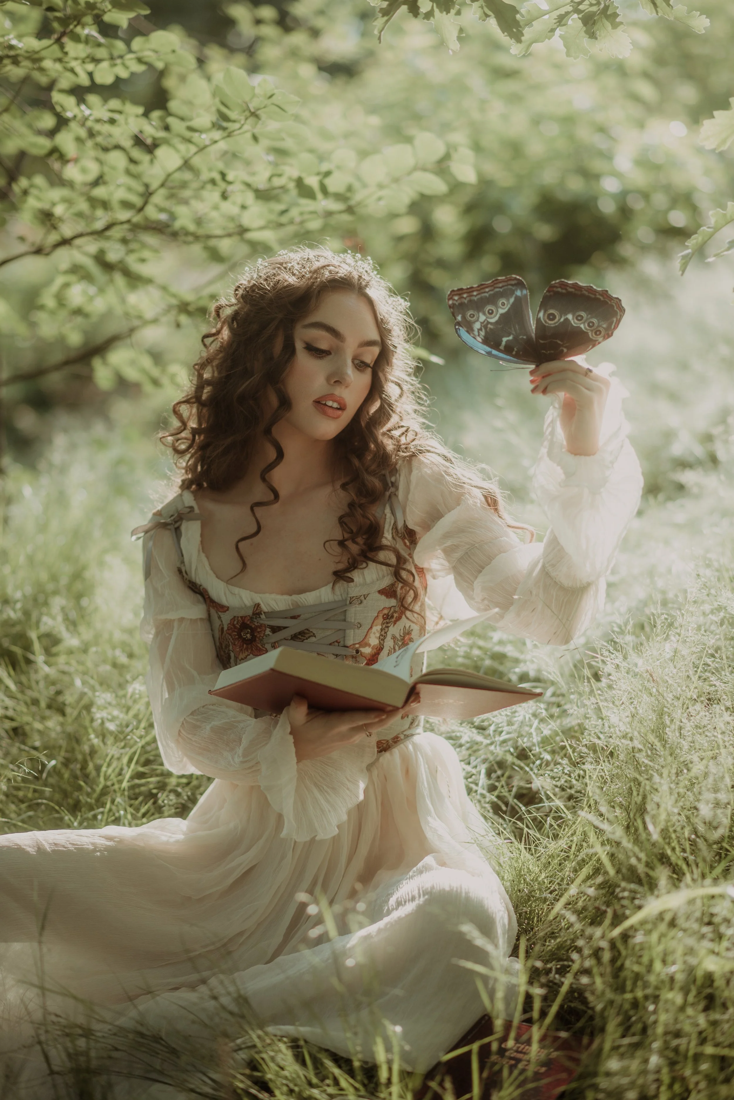 A woman with curly brown hair wearing a cream-colored romantic dress with embroidery sits in lush green grass, reading a book and holding a large butterfly-shaped butterfly net.