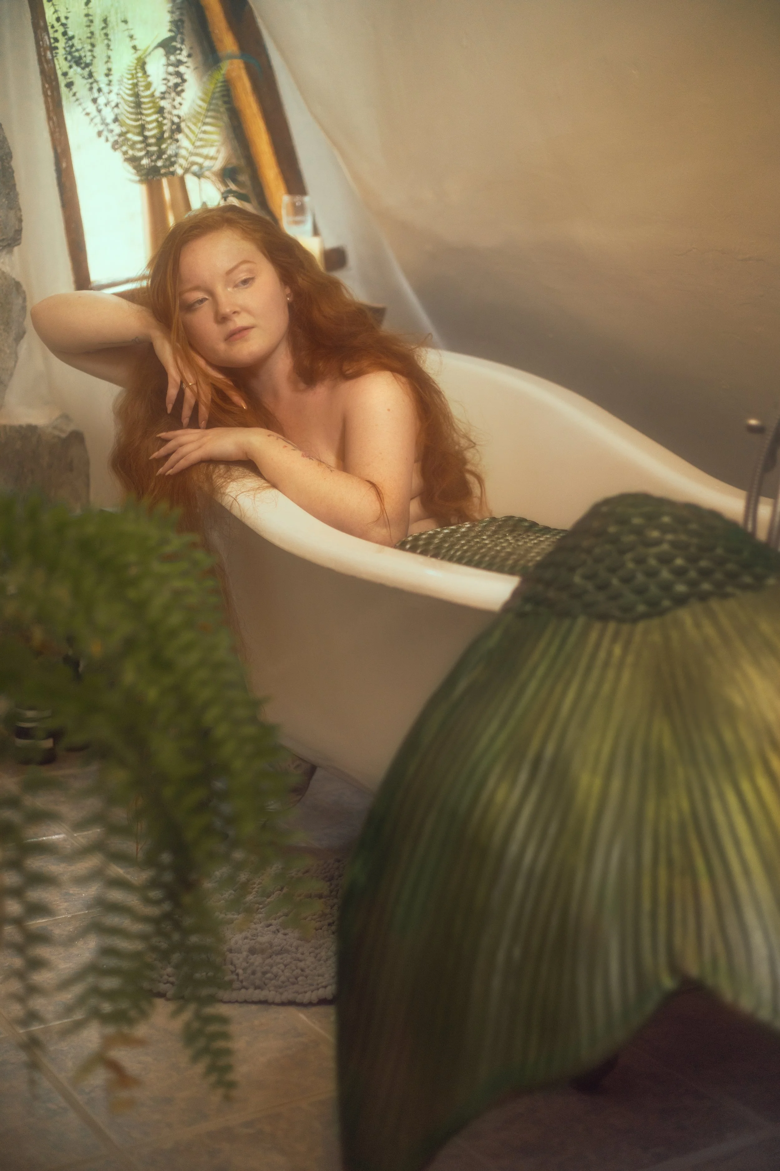 Red-haired woman sitting in a vintage clawfoot bathtub in a cozy, rustic room, surrounded by plants.