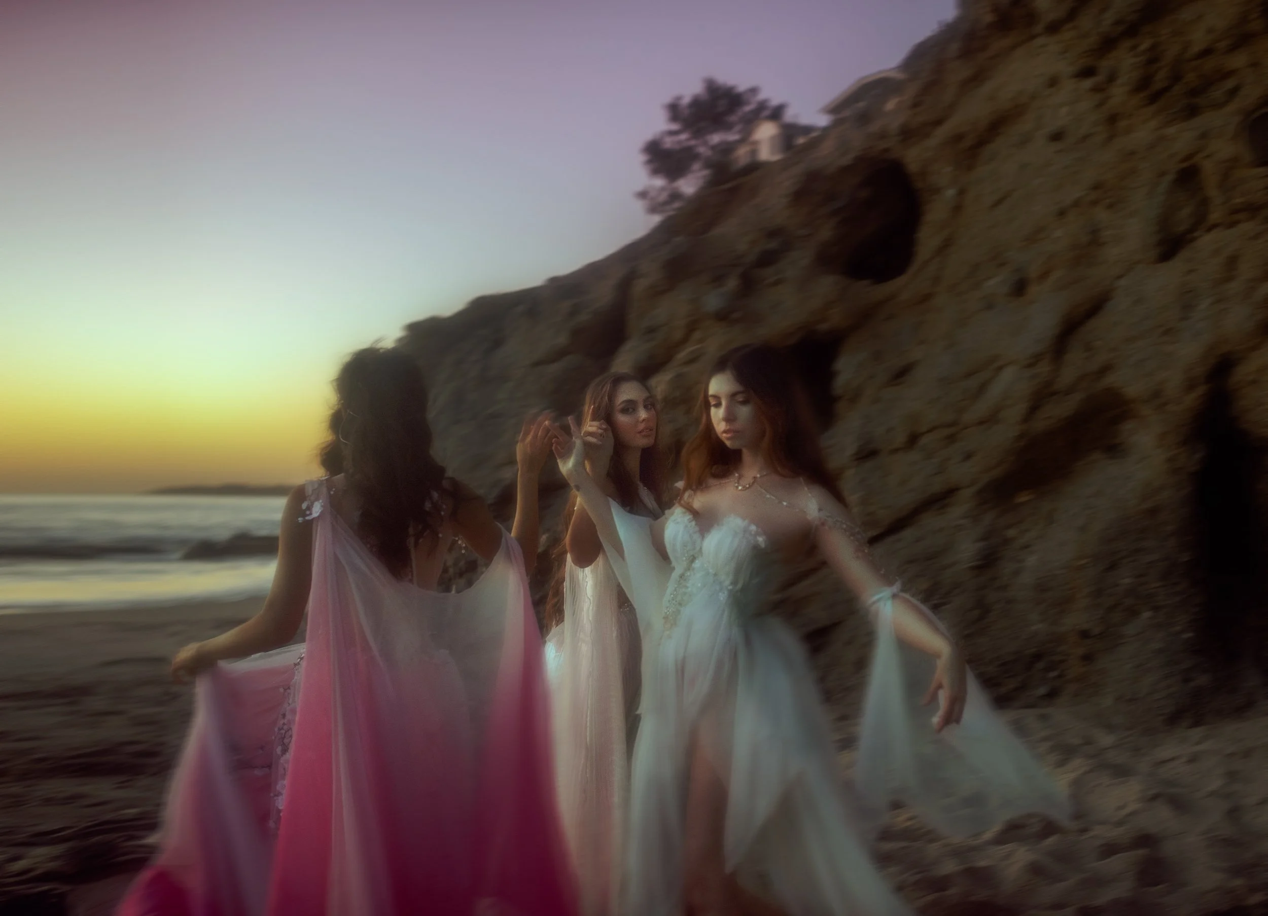 Three women in flowing dresses on a beach at sunset, with large rocks in the background.