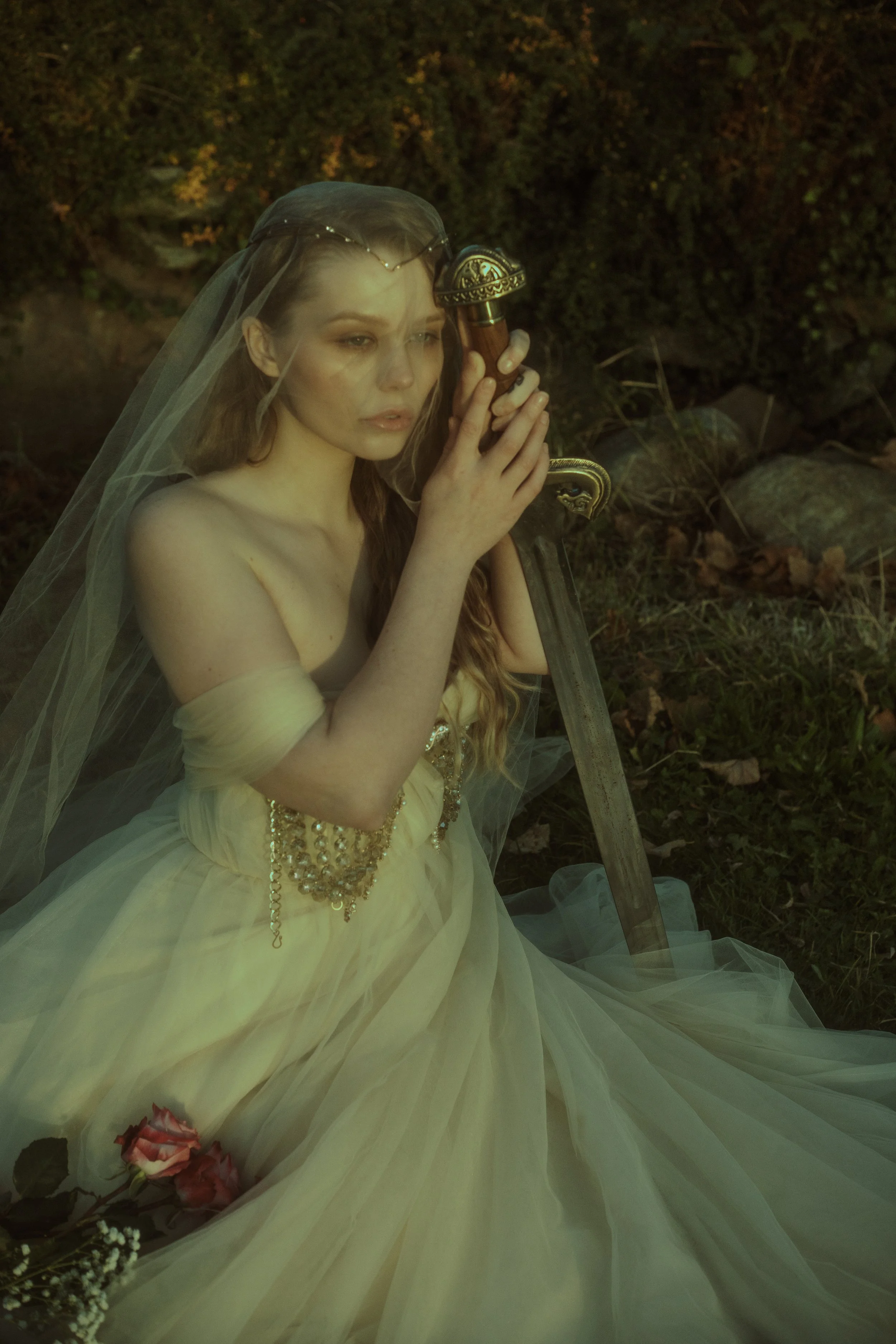 A woman in a yellow gown with a veil and tiara holding a sword in a forest-like outdoor setting.