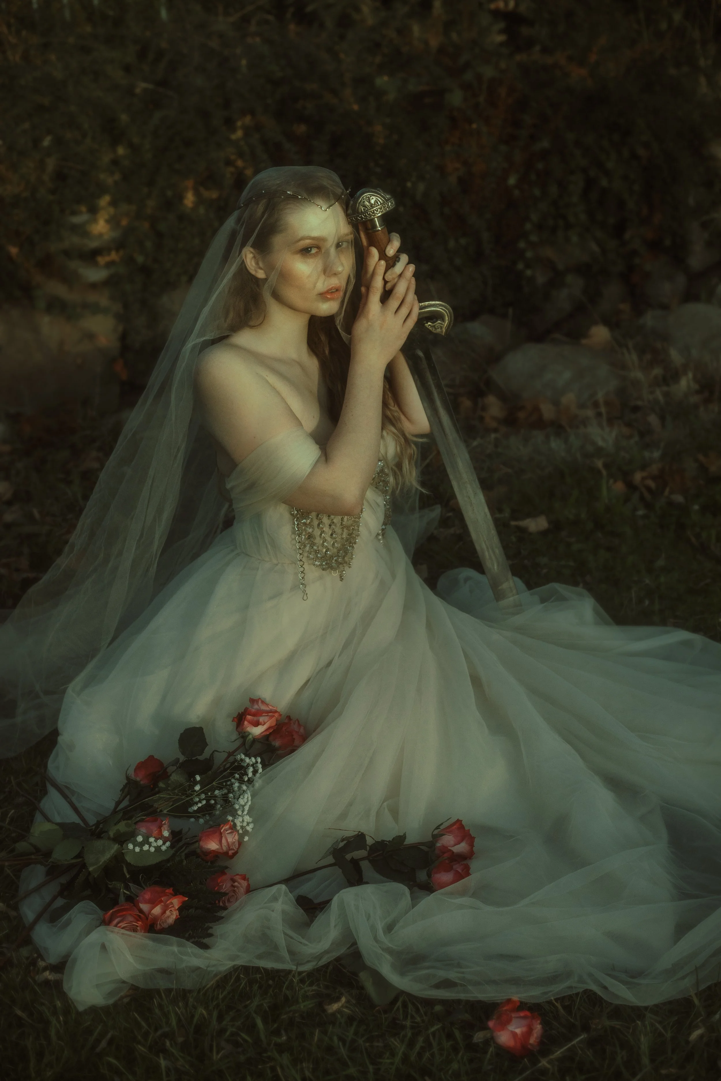 A woman dressed in a wedding gown with a veil, sitting on the grass outdoors at dusk, holding a sword vertically in front of her face, with pink roses and greenery around her.