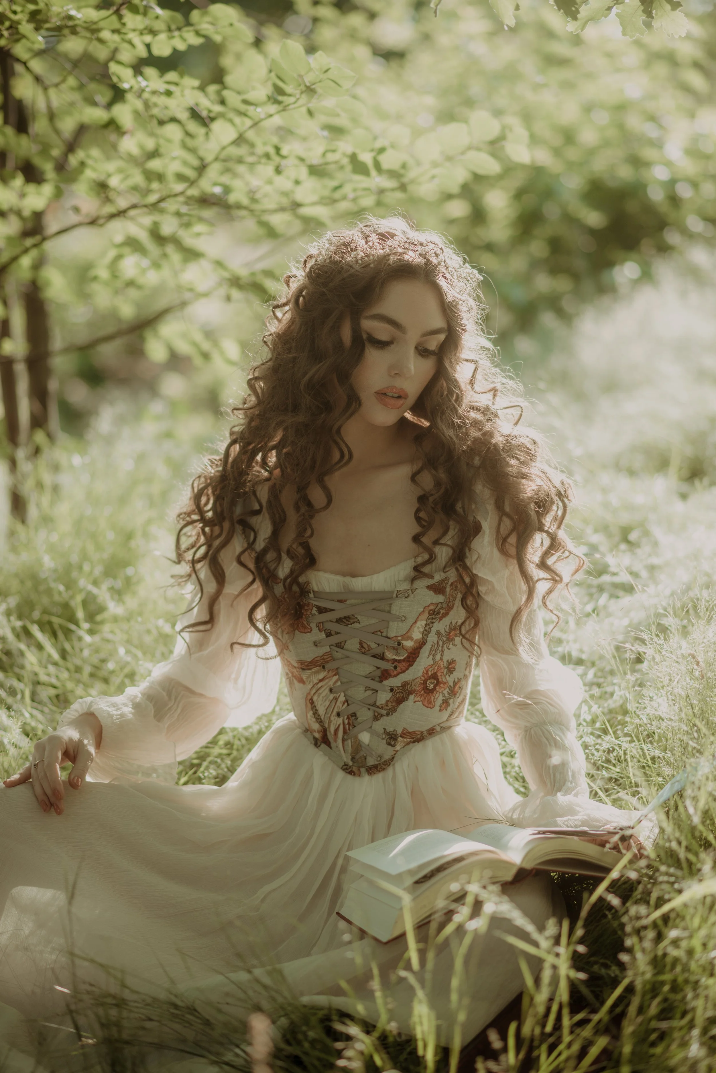 A woman with long, curly hair sits outdoors in a sunlit, green field, wearing a cream-colored dress with a detailed, lace-up bodice, looking down at an open book in her lap.