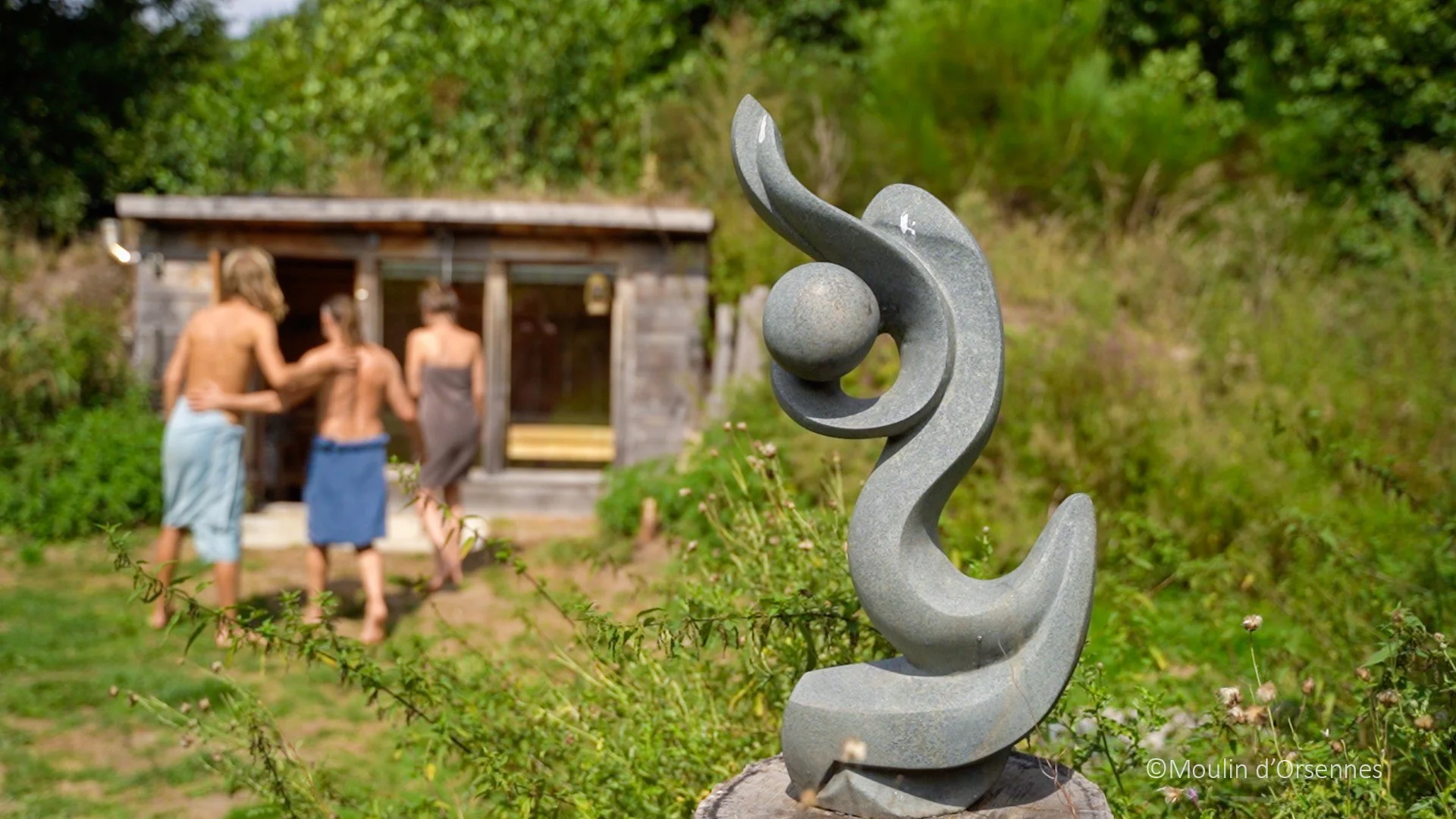 Een abstracte stenen sculptuur in de voorgrond met drie mensen die in de achtergrond voor een kleine houten hut staan.