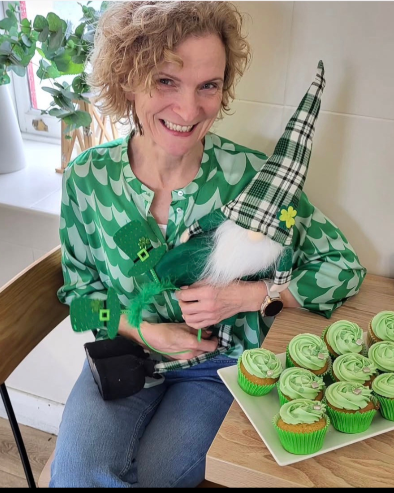 Woman in green patterned shirt holding a gnome decoration with a plaid hat, white beard, and green outfit, sitting next to a plate of green frosted cupcakes.