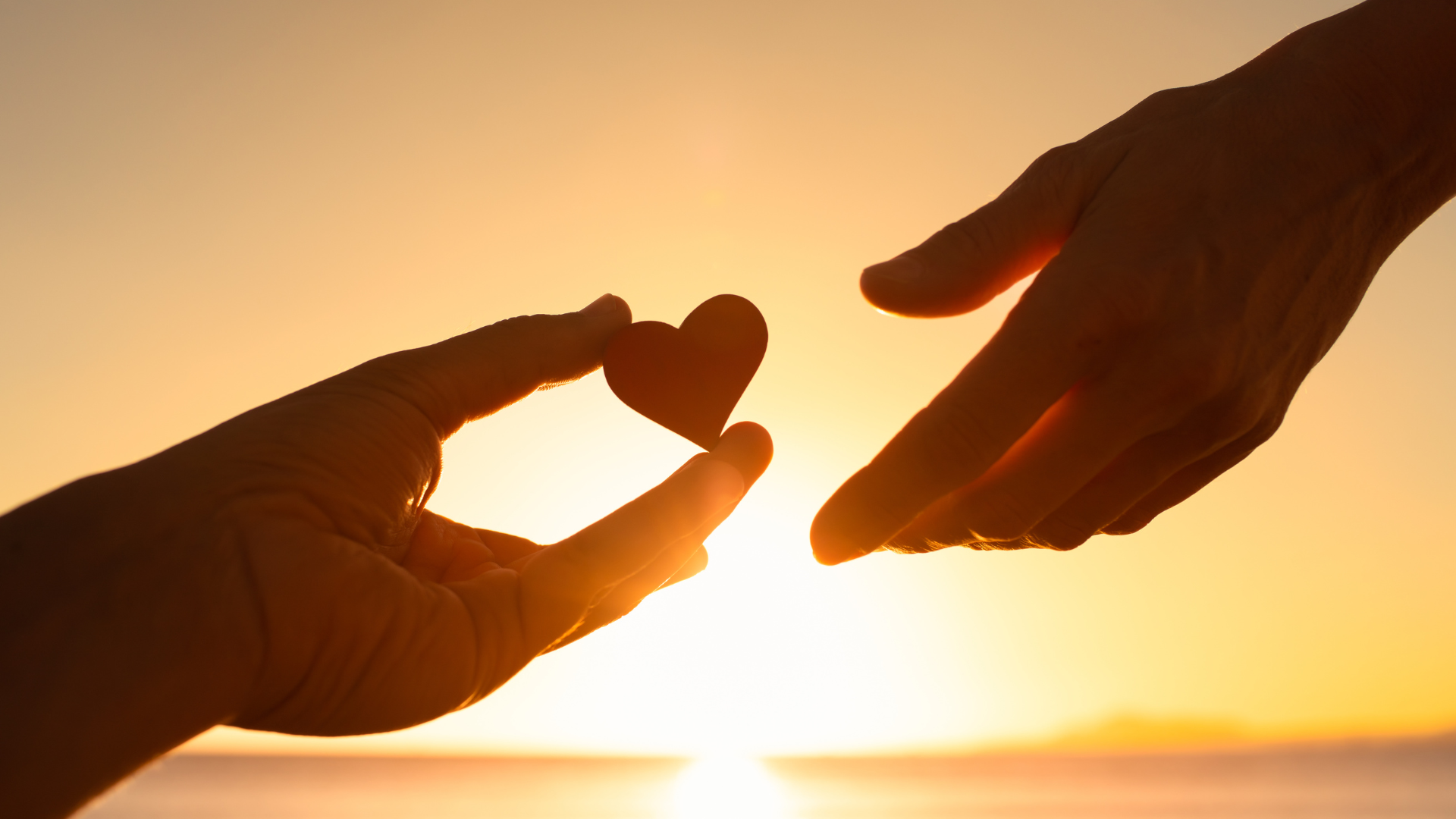 Two hands passing a heart-shaped symbol at sunset, representing generosity, compassion, and giving to others.