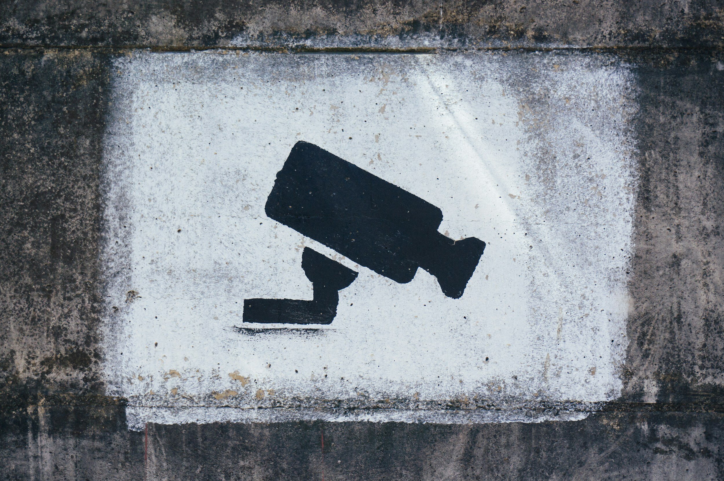 A stencil graffiti of a security camera painted in black on a white square background on a weathered concrete wall.