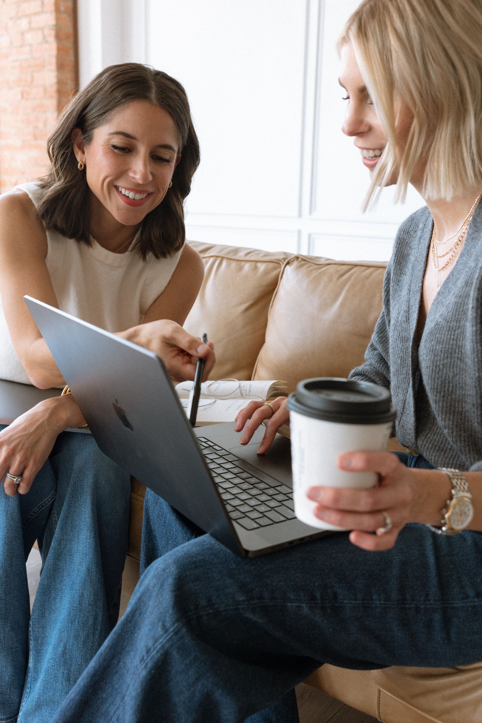 Two women sitting on a beige couch, smiling and looking at a laptop screen, while one holds a coffee cup in her hand.