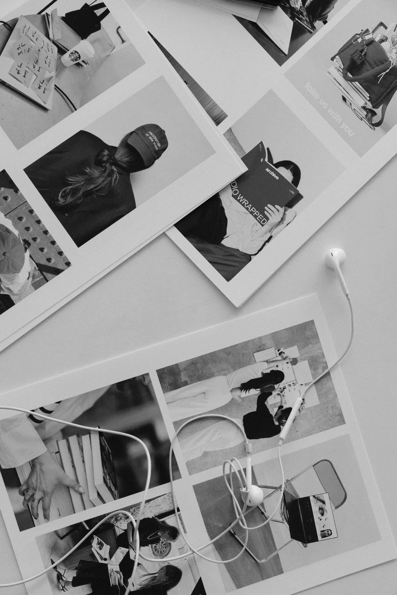 Black and white photograph of various pictures and notes spread on a table, with a white earphone on top, alongside electronic equipment and people in the background.