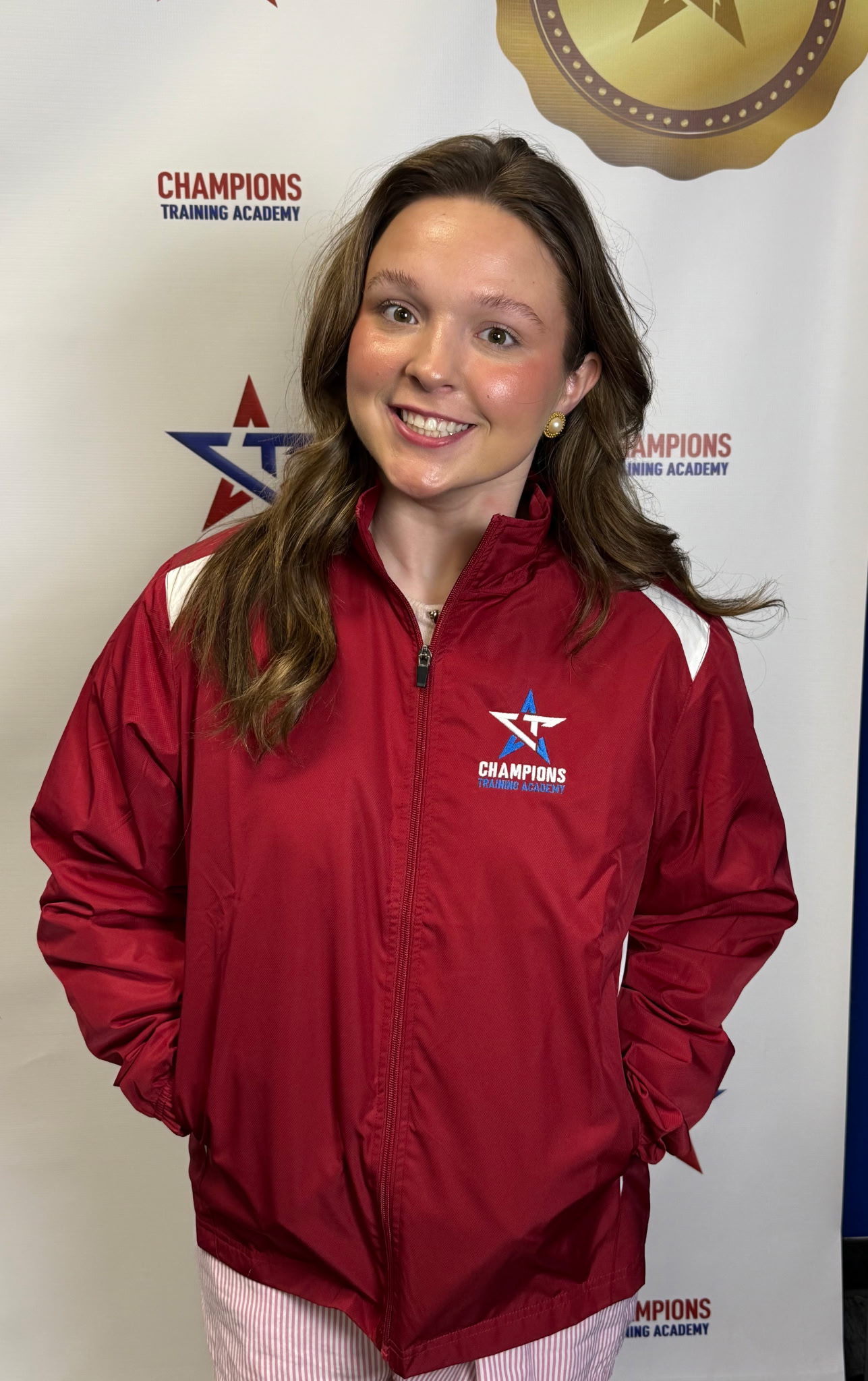 Young woman with long brown hair wearing a red Champions Training Academy jacket, smiling, standing in front of a white background with Champions Training Academy logos.