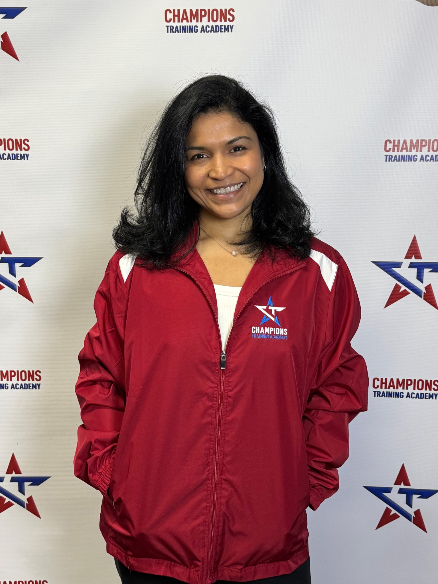 Smiling woman with black hair in a red jacket with 'Champions Training Academy' logo, standing in front of a white background with the same logo repeated.