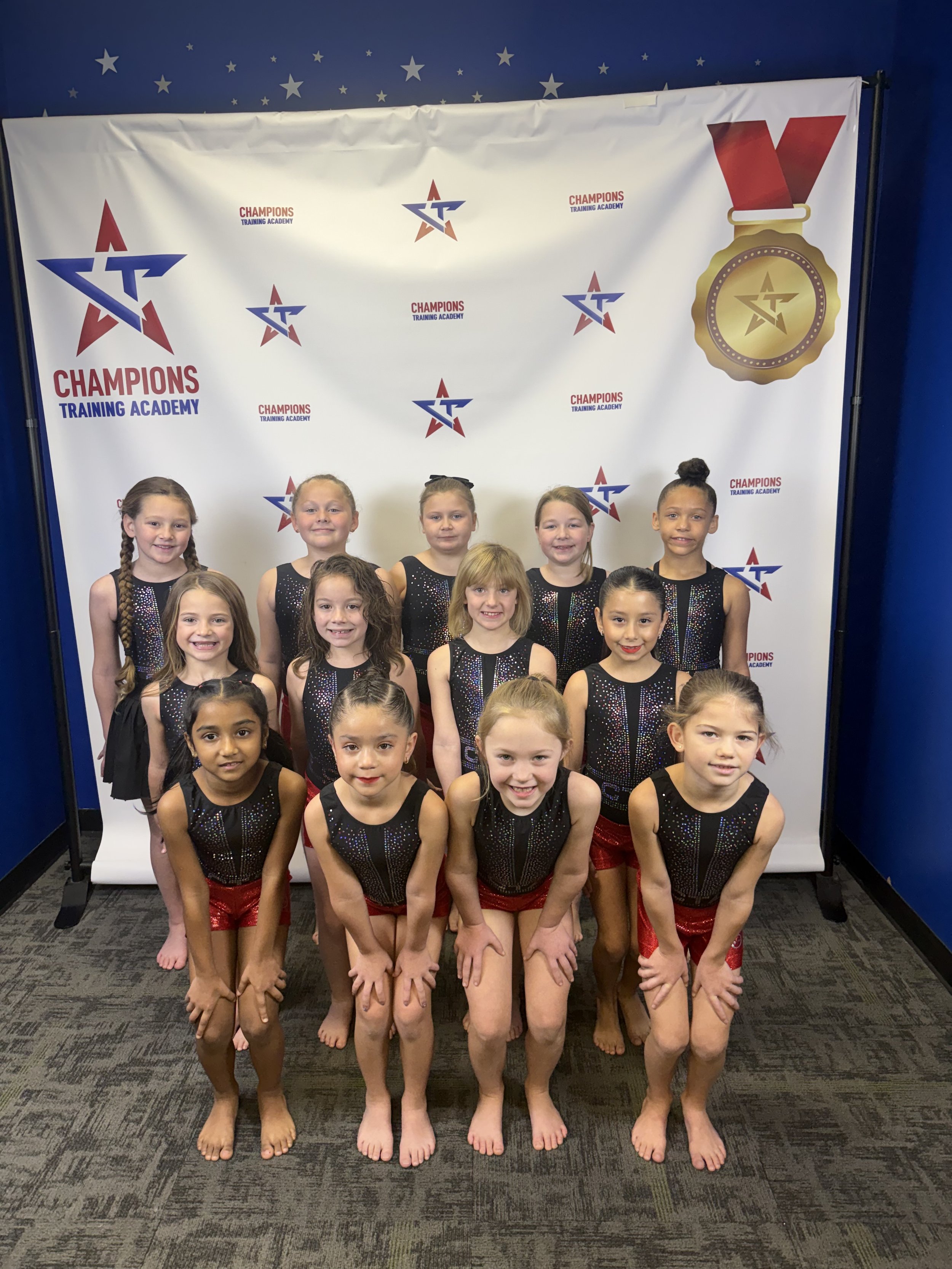 Group of seven young girls in navy blue gymnastics leotards with rainbow designs, standing confidently with arms crossed, against a colorful smoky backdrop, promoting the USAG Pre-Team for 2023-2024 at Champions Training Academy.