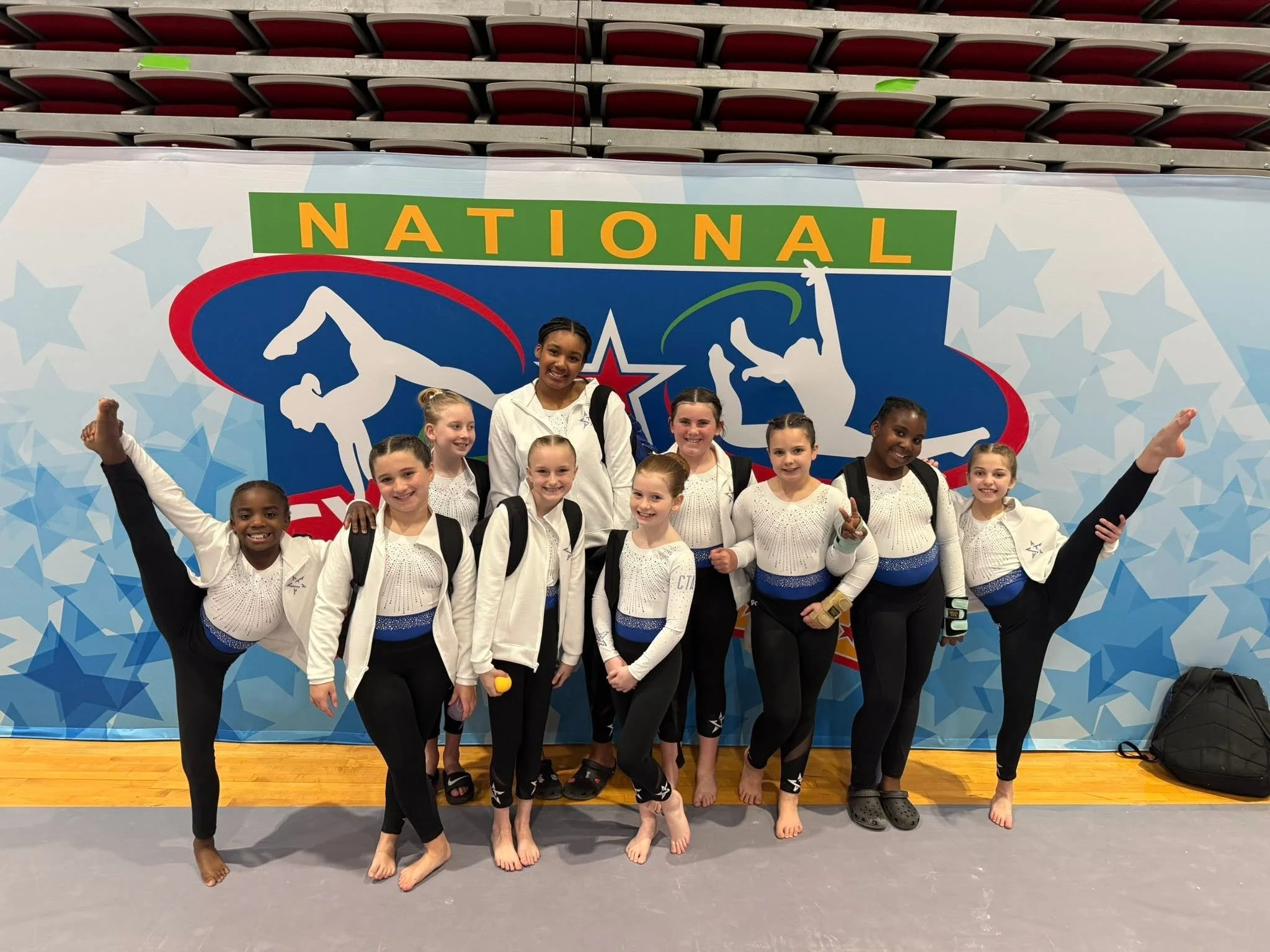 Five young girls in red and black gymnastics leotards sitting on the floor, smiling, with a backdrop that reads "Champions Training Academy" and confetti falling around them.
