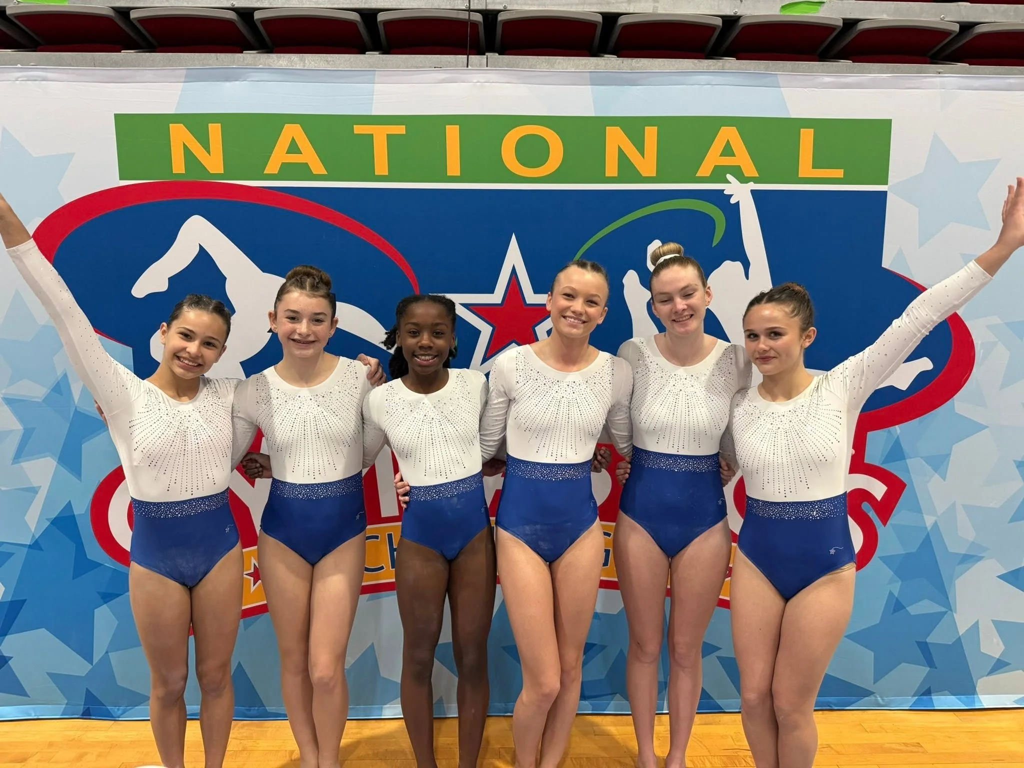 Five young female gymnasts in red and navy blue uniforms posing confidently against a colorful background with pink, blue, and purple hues. The text indicates their team status as 'USAG Level 4' for the 2023-2024 season, associated with Champions Training Academy.