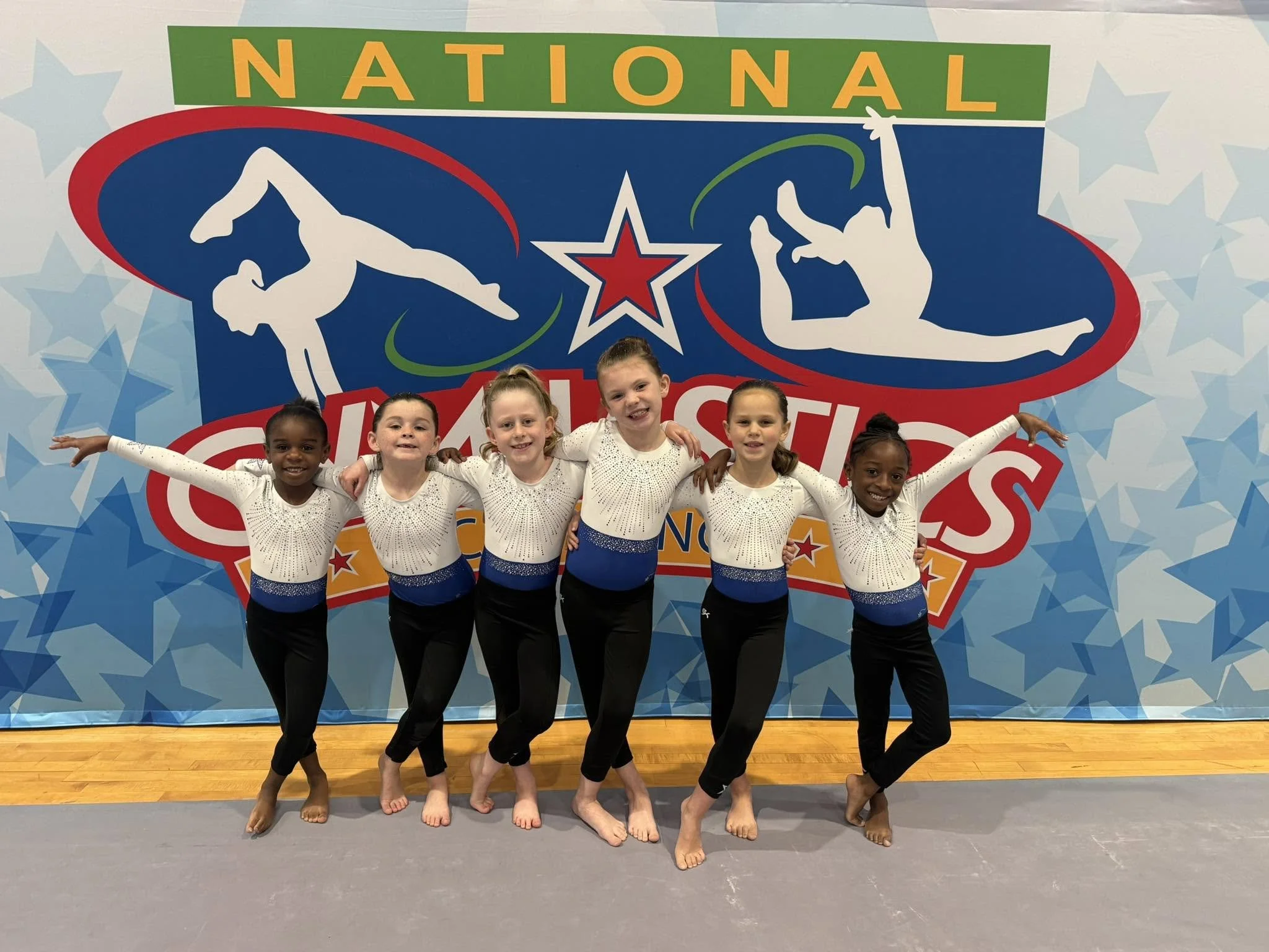 Group of cheerleaders in red and black uniforms posing on stage with a large display that reads "Champions Training Academy 2024-2025" and a confetti background.
