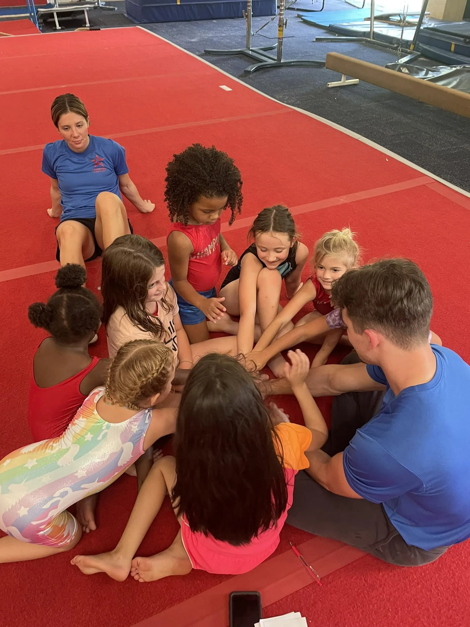 Children and a coach sitting in a circle on a red gym floor, raising their hands, with a blue and white gymnastics mat in the background.