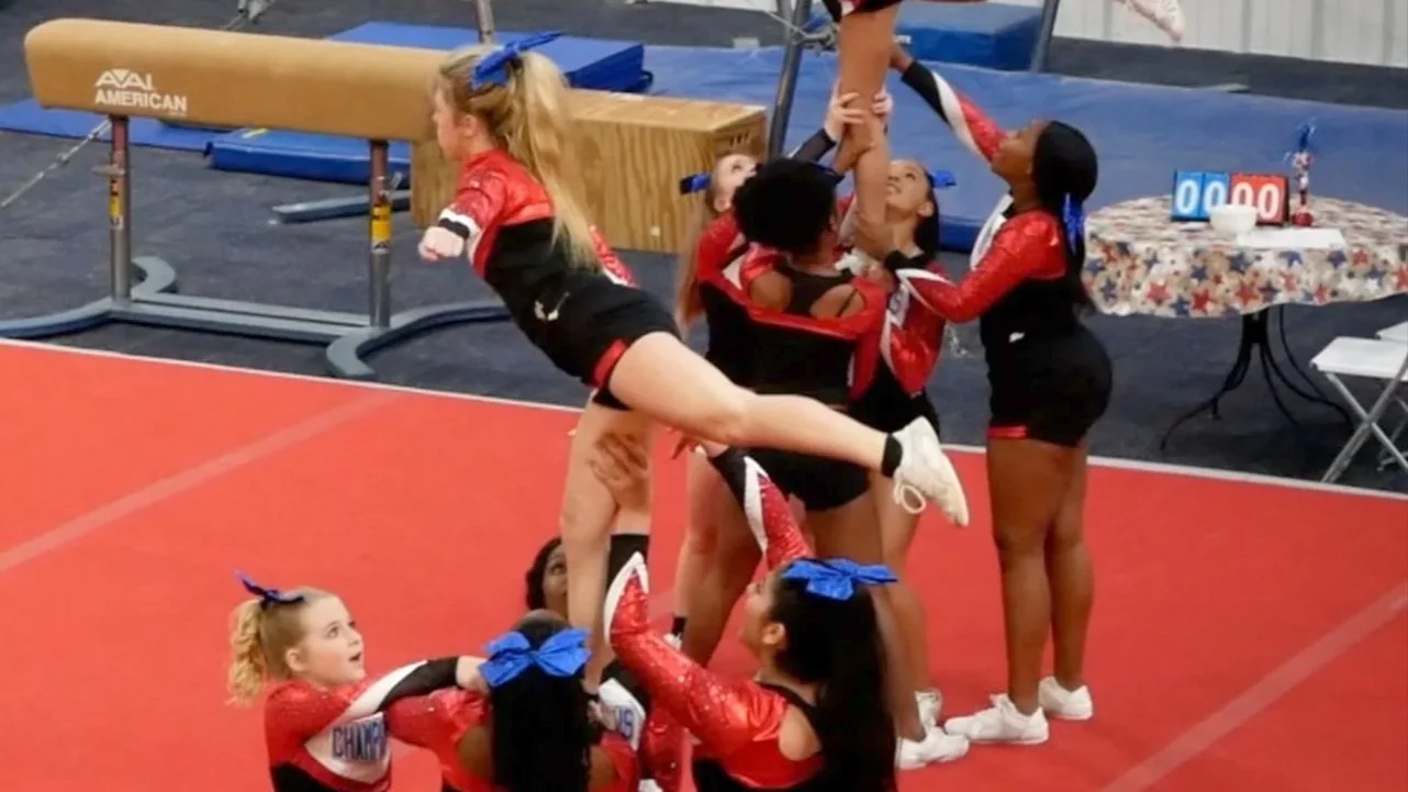 Cheerleaders performing a stunt on a gymnastics mat at a cheerleading competition, with a cheerleading table and score banners in the background.