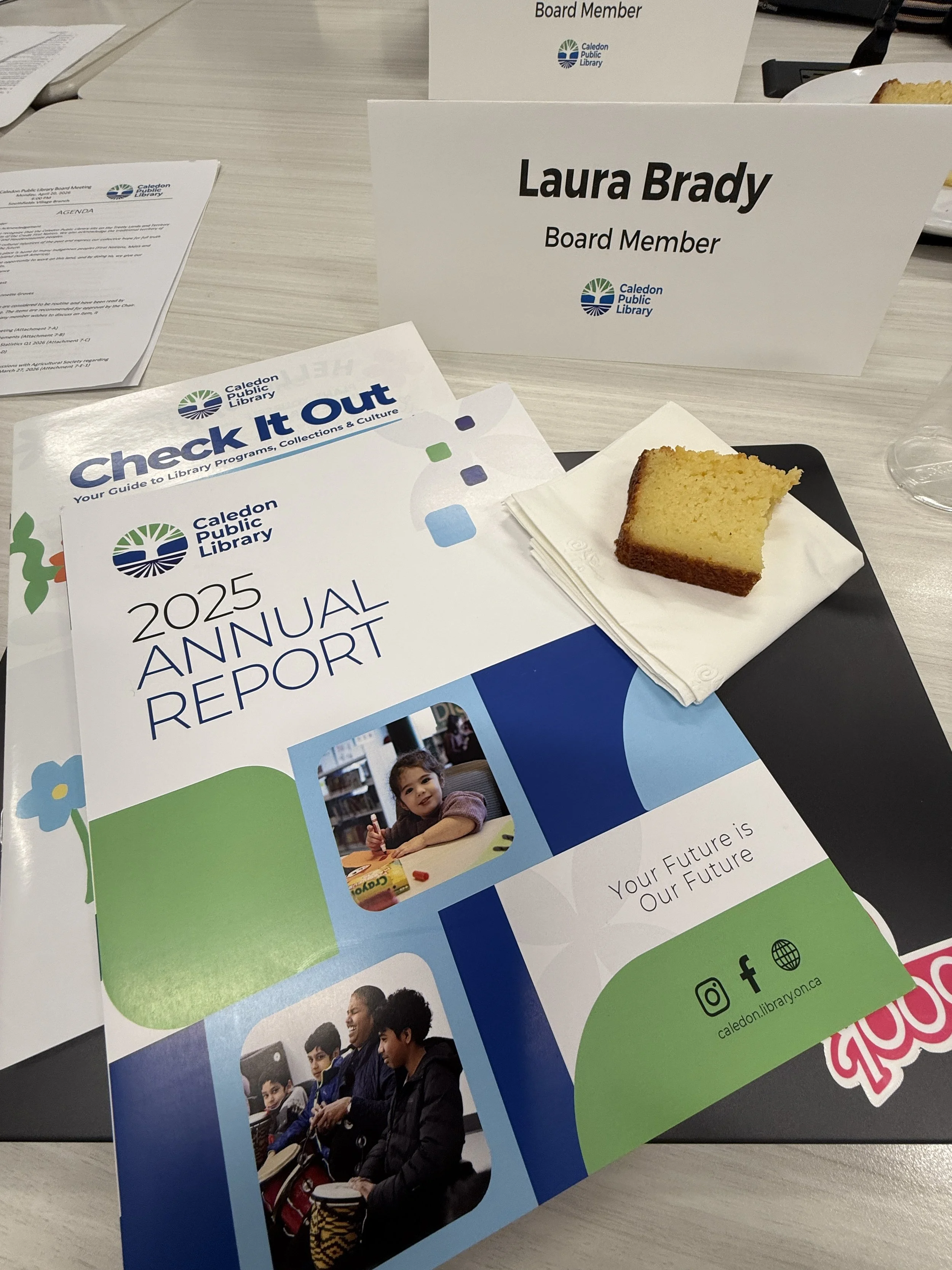 A piece of lemon loaf on a napkin next to the Caledon Public Library's 2025 Annual Report, and below a name card reading, Laura Brady, Board Member.