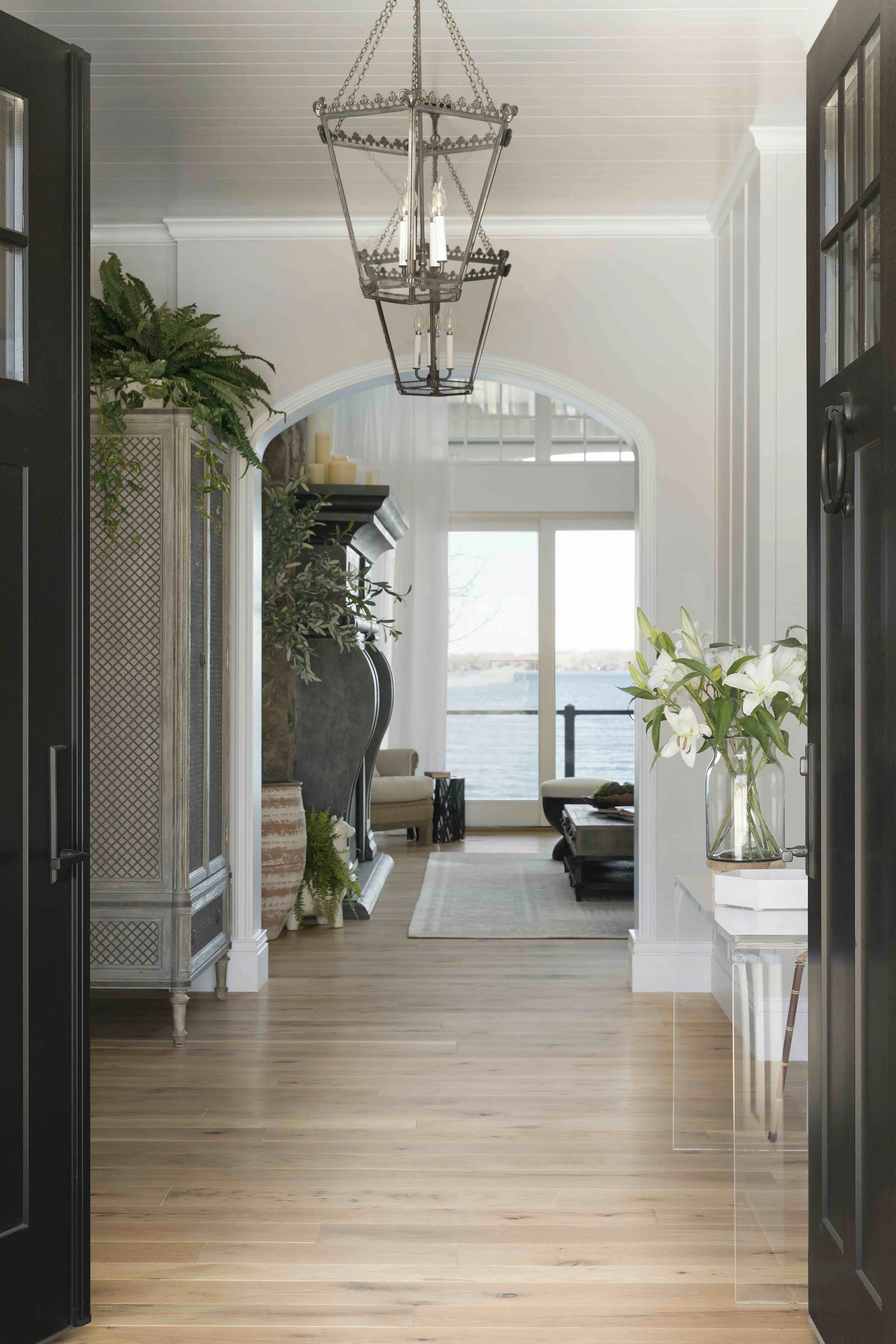 A view through a double door into an entryway with wood floors, large pendant lights and a tall cabinet.