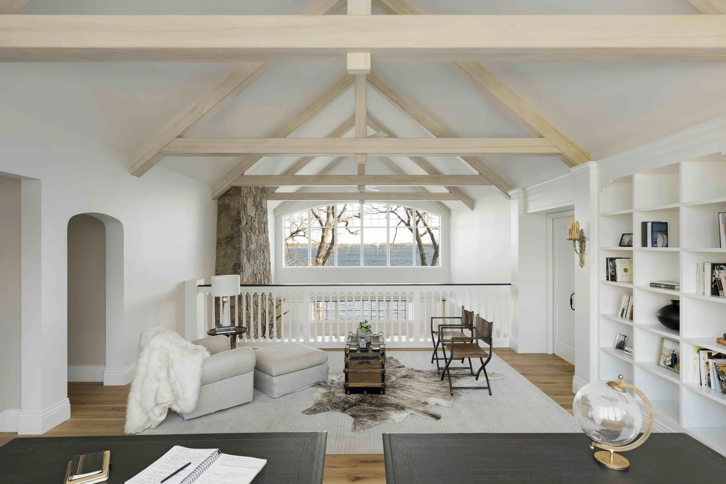 A loft sitting area with light wood ceiling beams, white built-in book shelves and large window overlooking water