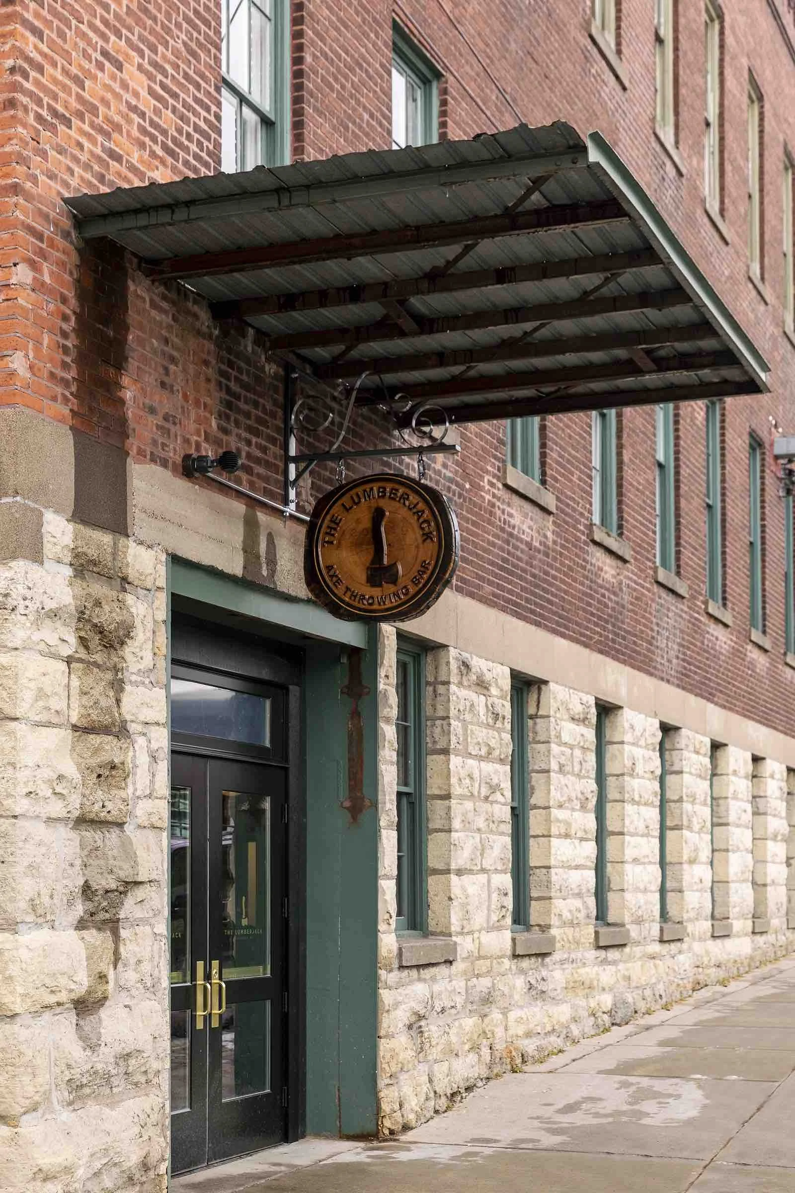 A brick and stone building with a green doorway and tree slice hanging sign with The Lumberjack Axe Throwing Bar logo