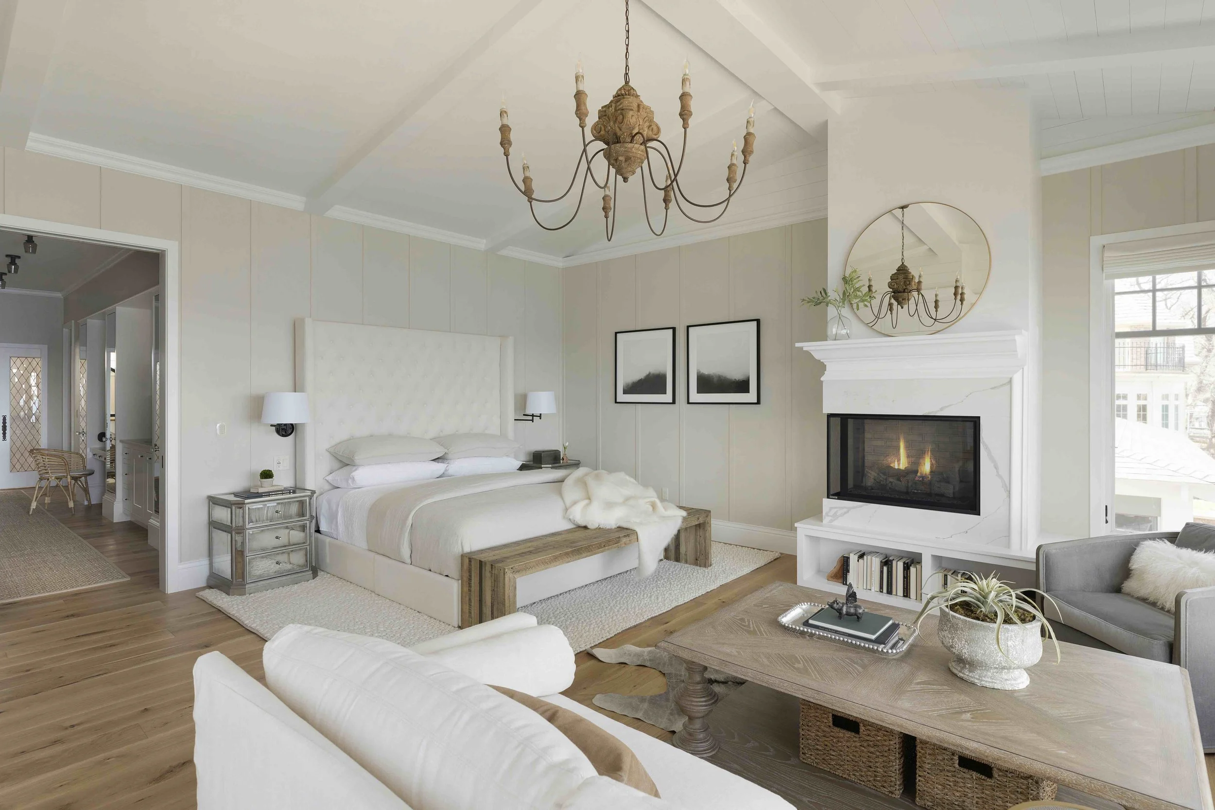 A large white master bedroom with light wood floors, light walls, white bedding, large chandelier, sitting area and gas fireplace