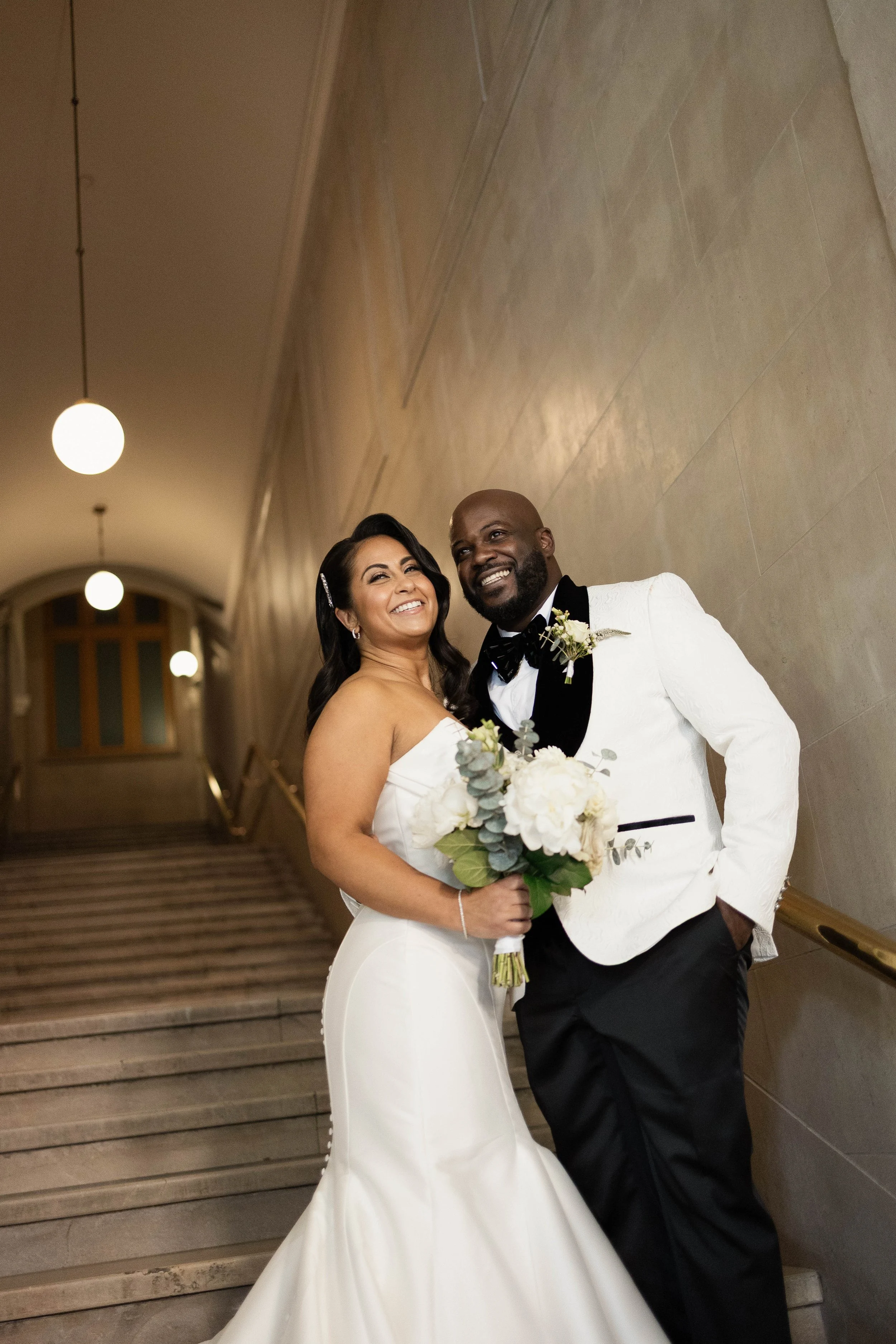 a bride and groom stand together laughing in a stone stairwell. There are rounded light fixture hanging along the ceiling.