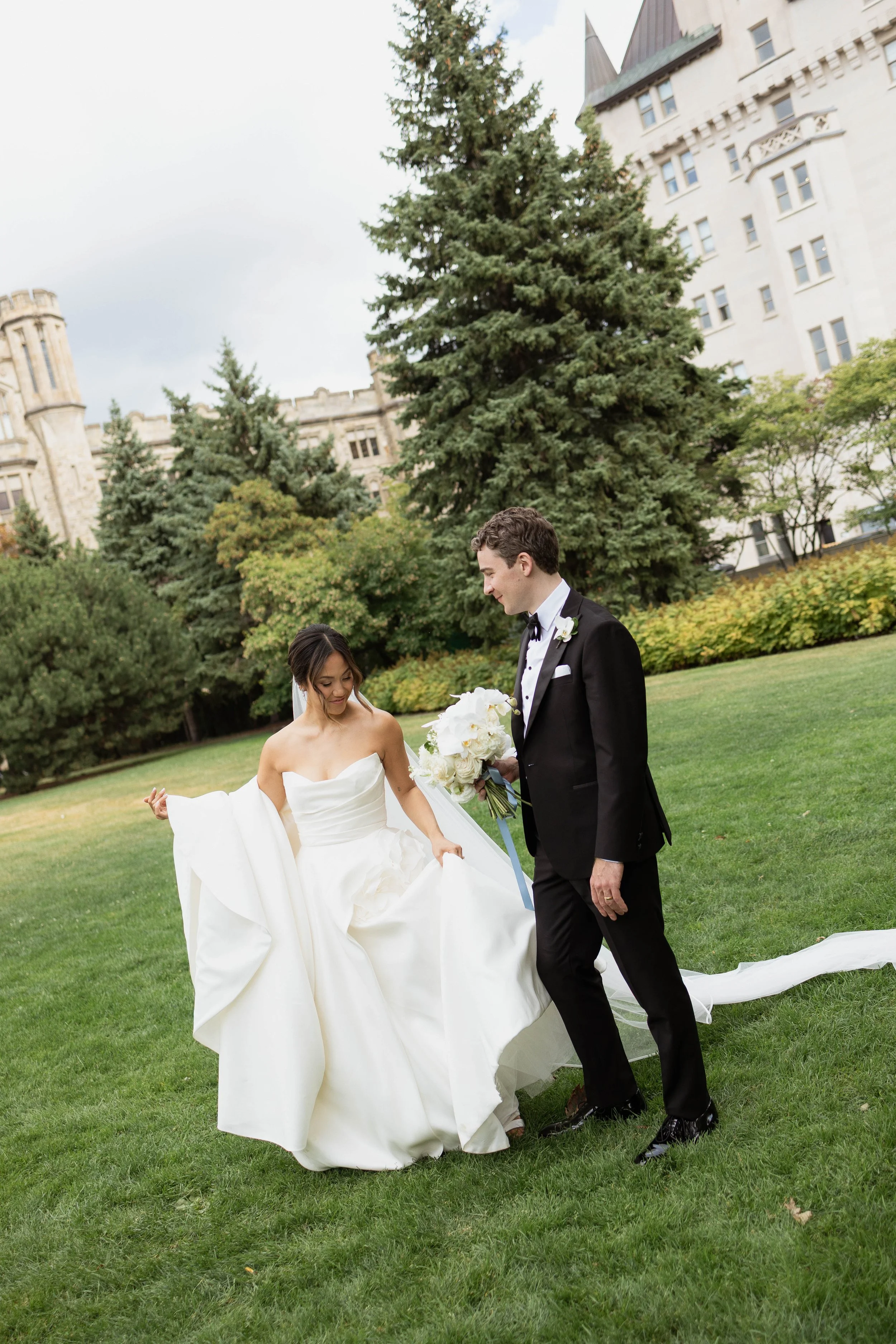 A full-body view of a bride and groom walking side by side. They are outside on the grass, with a church in the background obscured by trees.