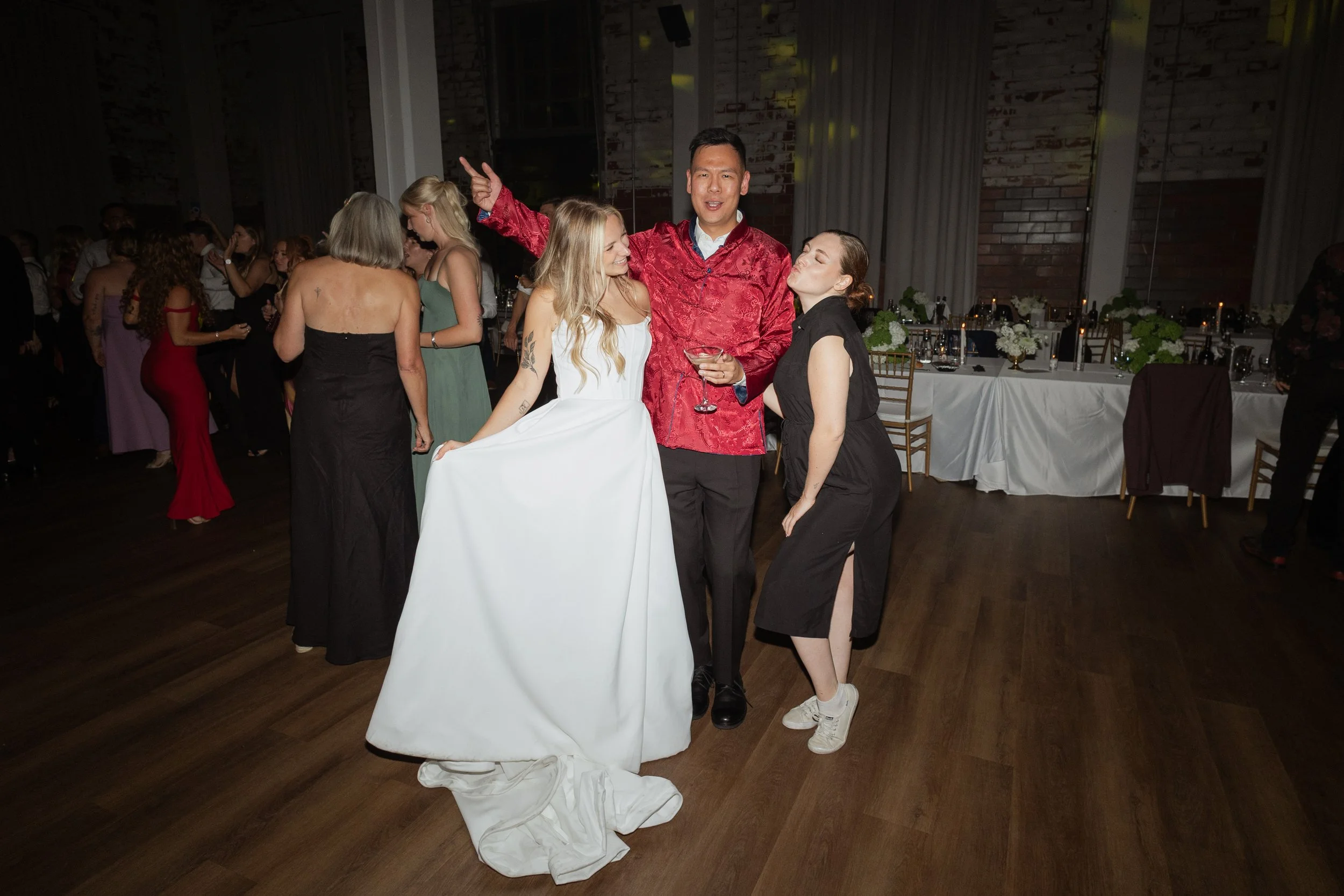 Sam stands with a bride and groom at their indoor reception. They are all laughing together.