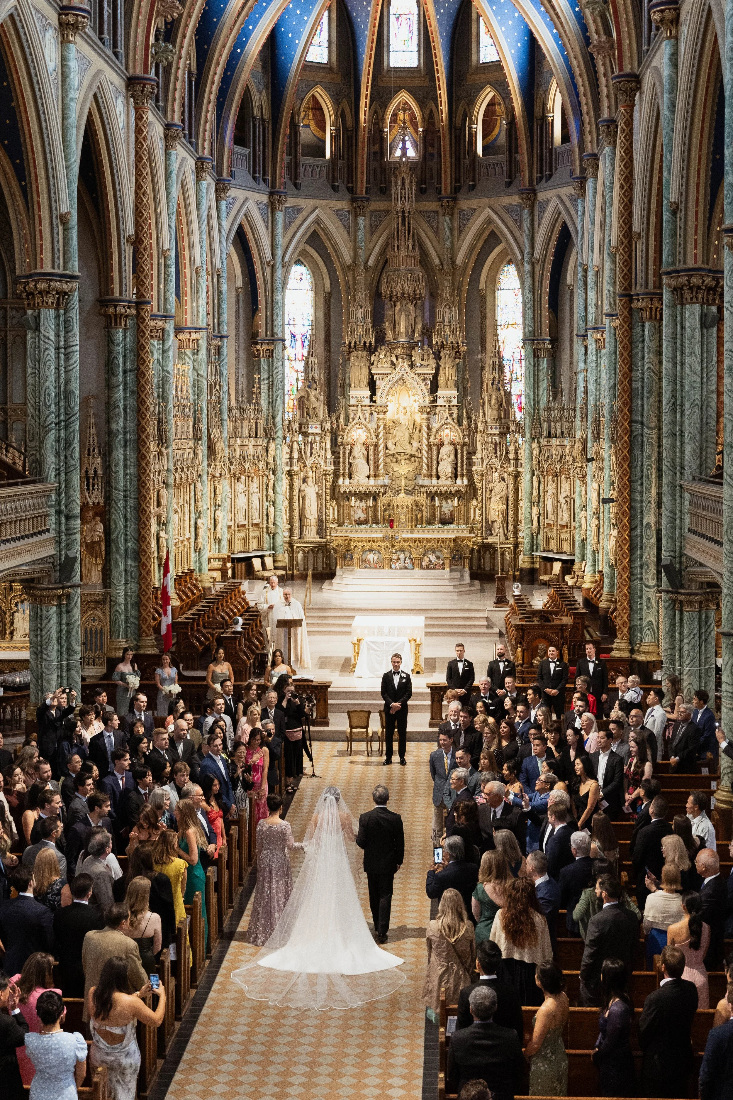 taken from above, a bride walks down the aisle of a church to meet her groom at the altar. The pews are filled with people.