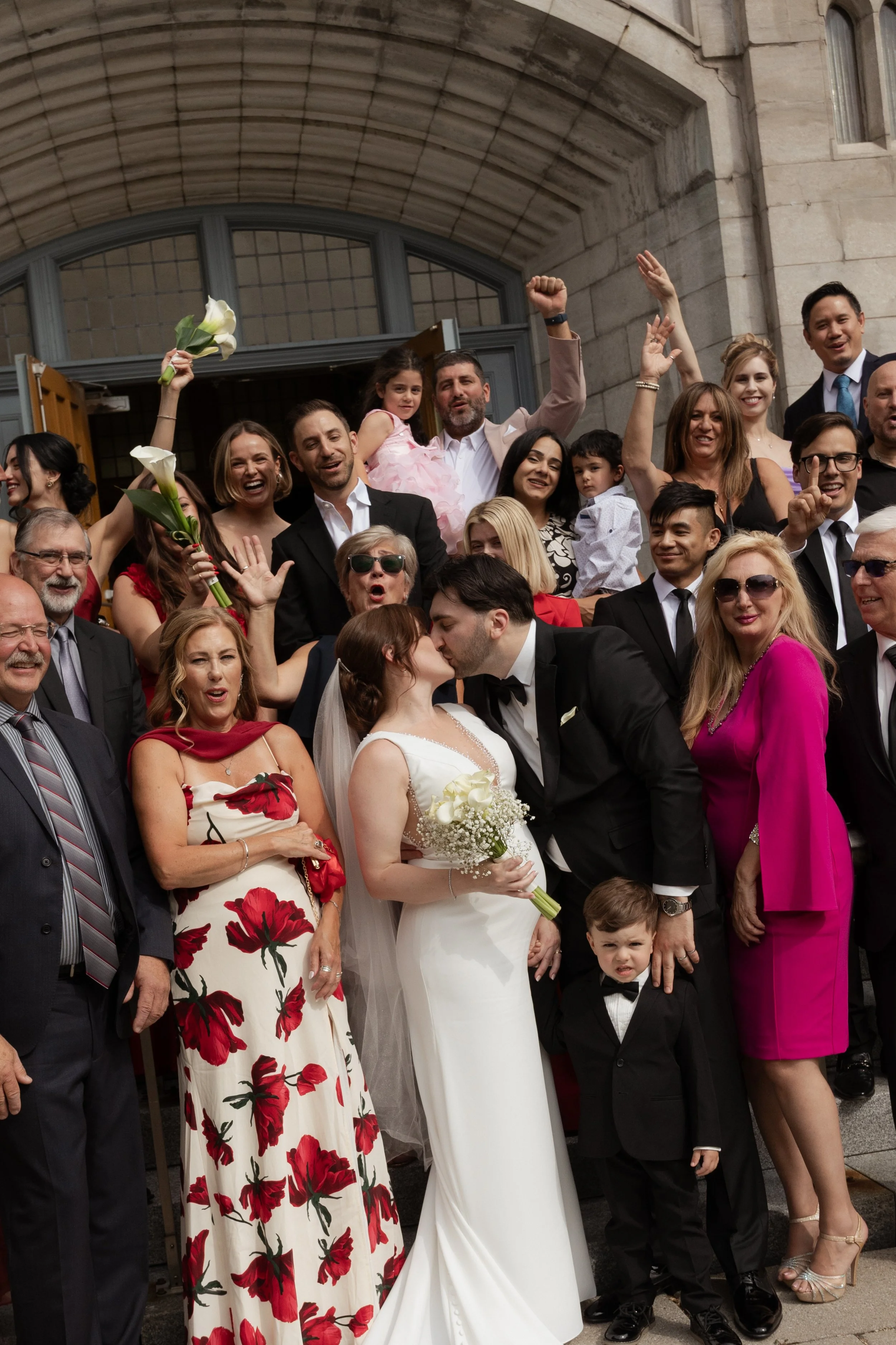 A newlywed couple stands on the steps of the church outside with all their guests standing around them. The guests cheer as the couple shares a kiss.