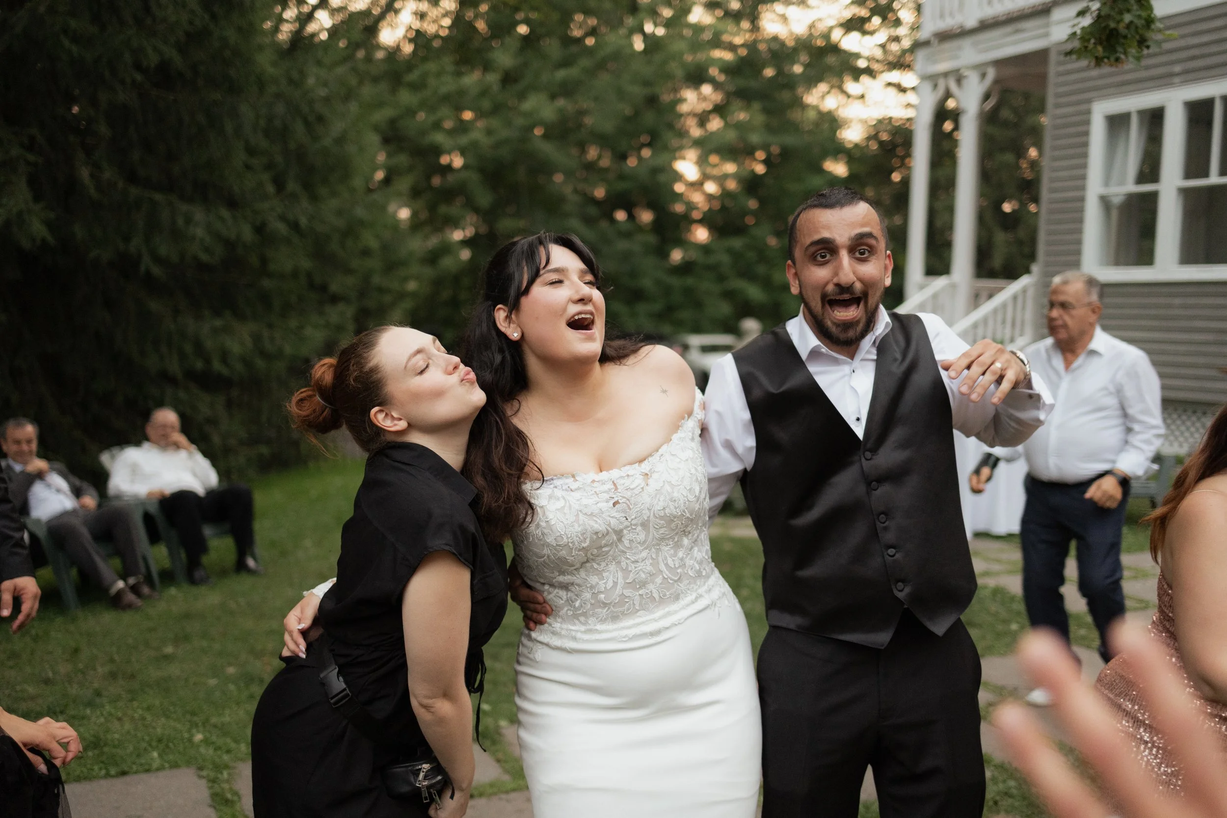 Sam stands with a bride and groom at their outdoor reception. She's blowing a kiss to them while they laugh
