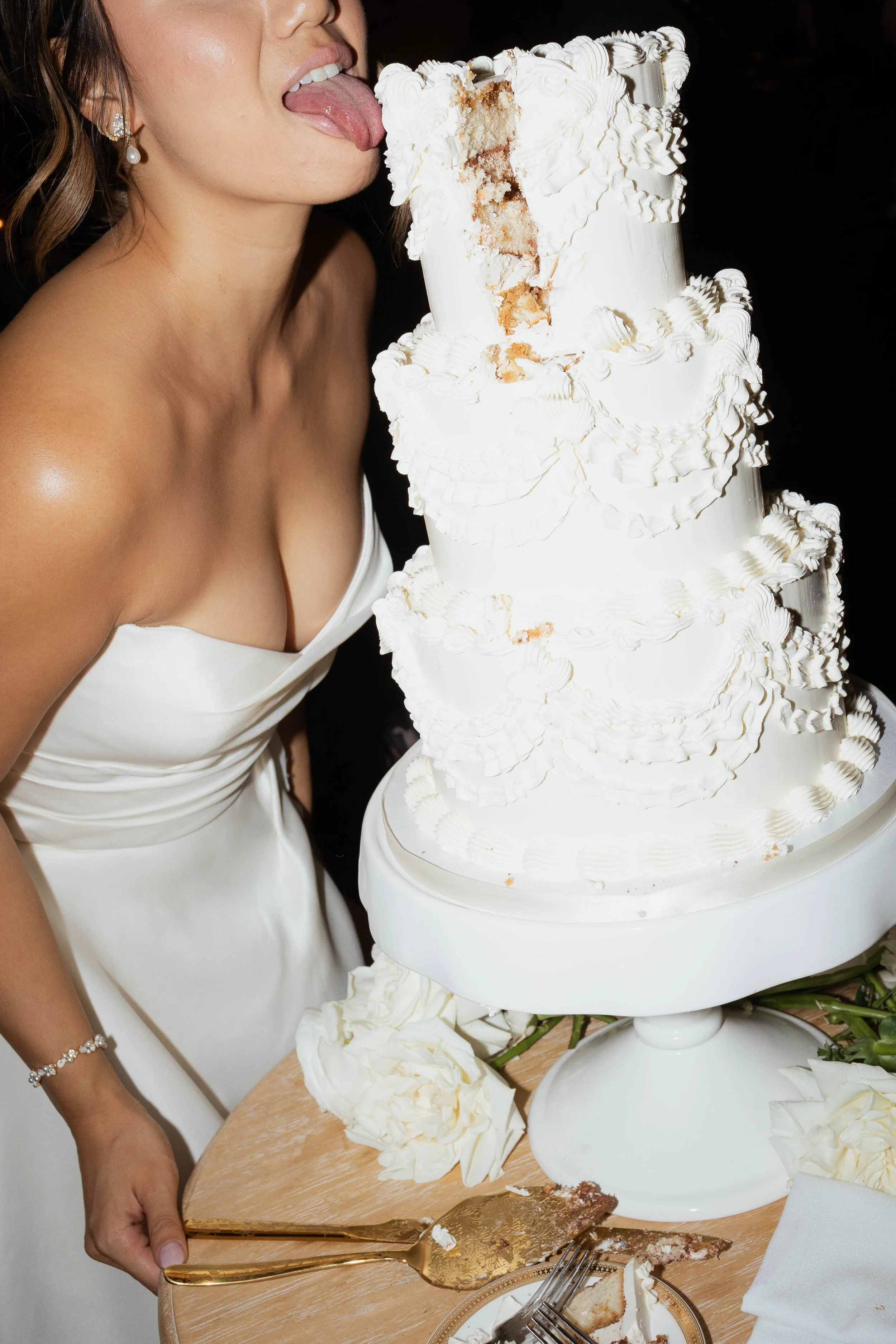 A flash photography image of a wedding cake with a slice taken out of the top tier. A bride stands next to the cake, pretending to lick it.