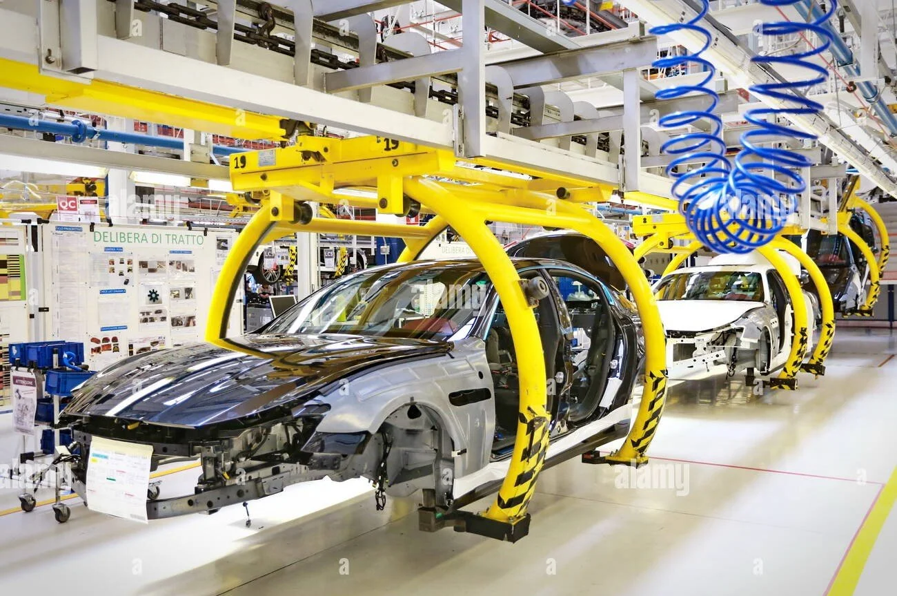 Automobile assembly line with partially assembled cars in a factory, featuring robotic arms, yellow safety frames, and blue air hoses.