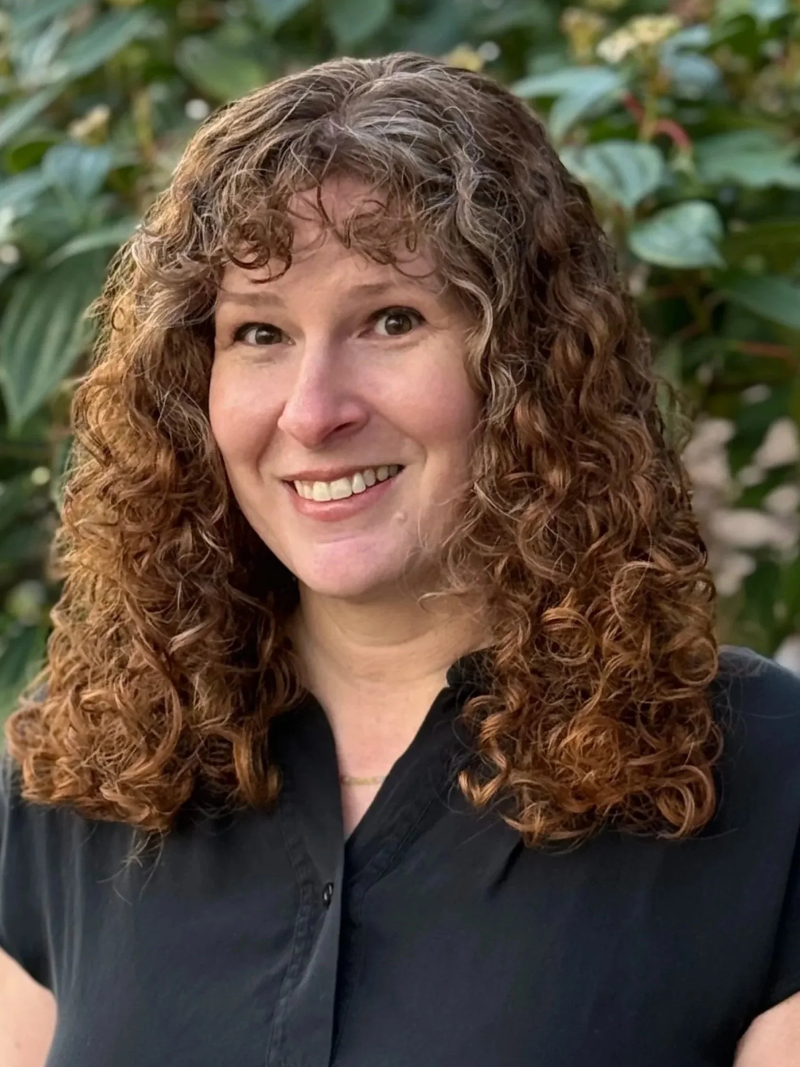 A woman with long, curly, reddish-brown hair smiling outdoors in front of green foliage, wearing a black shirt.